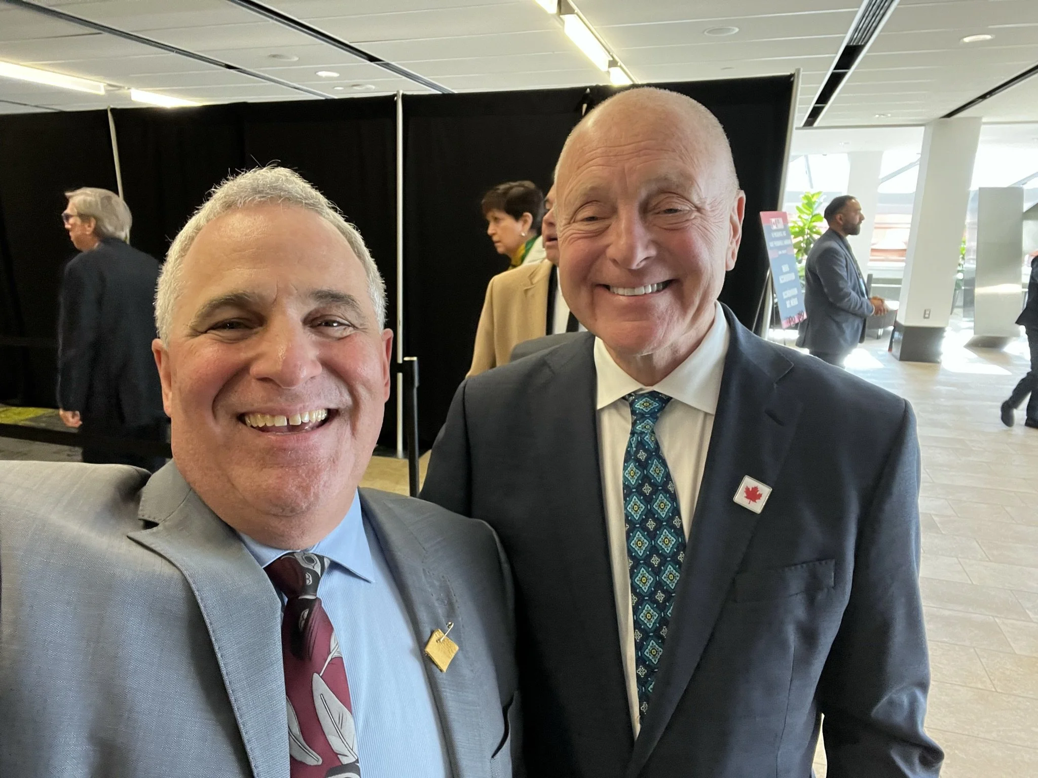 Two men in suits smiling for a photo at an indoor event, with other people in the background.