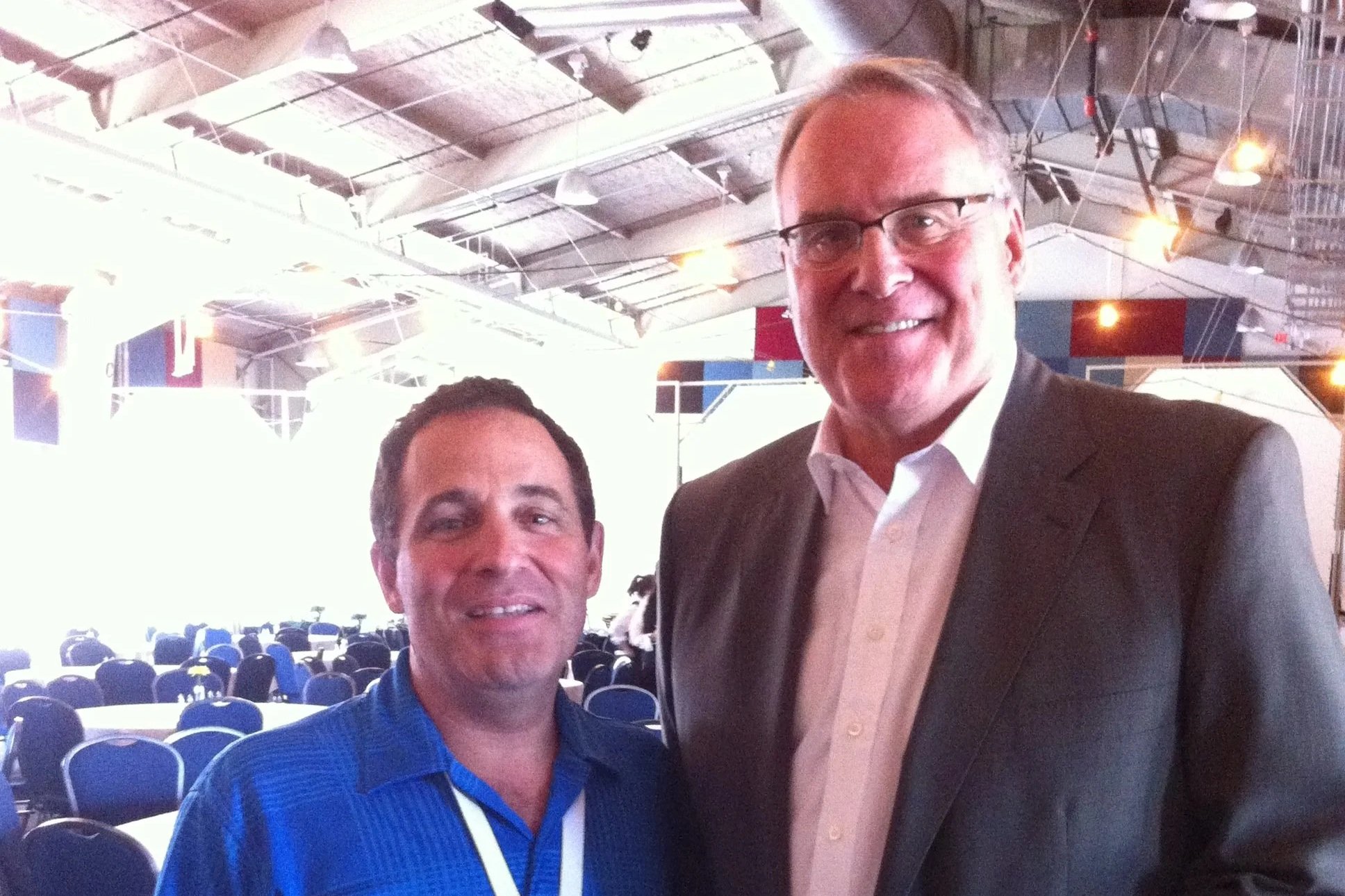 Two men smiling indoors at a conference or event, with rows of chairs and flags in the background.