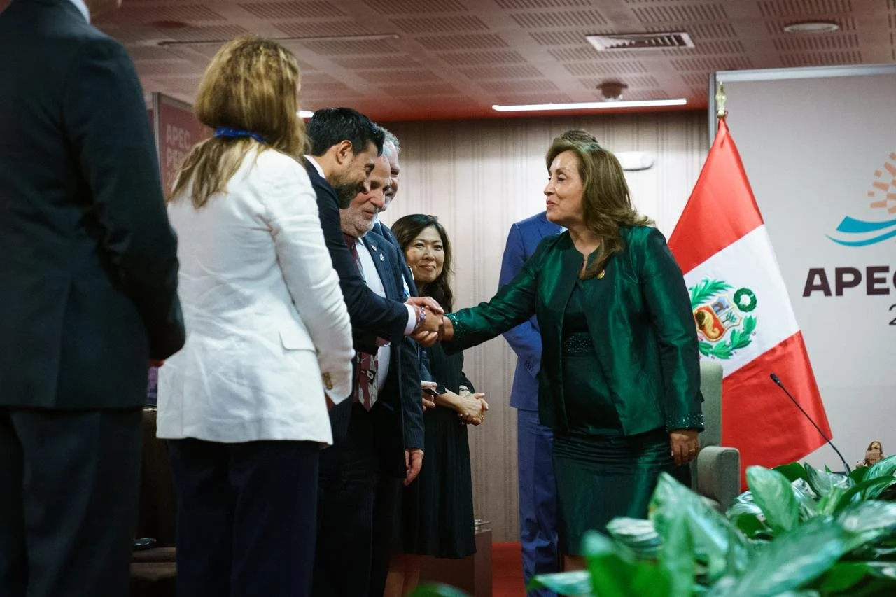 A woman in a green suit shaking hands with a man in a dark suit during a formal event, with other officials present in the background, Peruvian flag, and APEC banner.