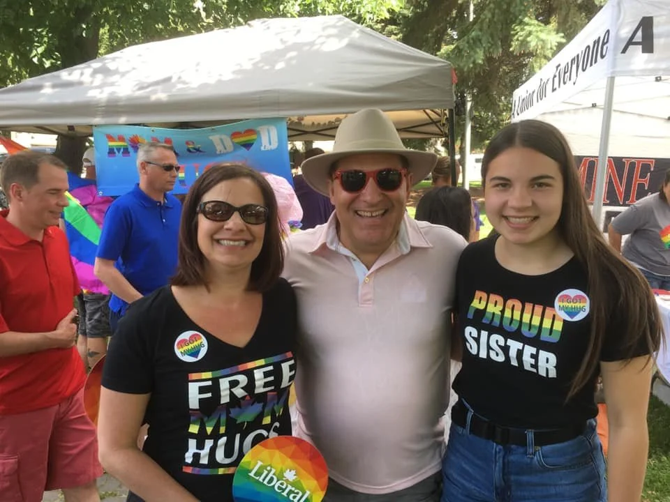 Three people smiling at an outdoor pride event, with two women and one man. The women wear black shirts with rainbow-colored text, one says 'FREE HUGS' and the other says 'PROUD SISTER.' The man in the middle wears sunglasses, a hat, and a light-colored shirt. They are in front of tents and other attendees, with trees in the background.