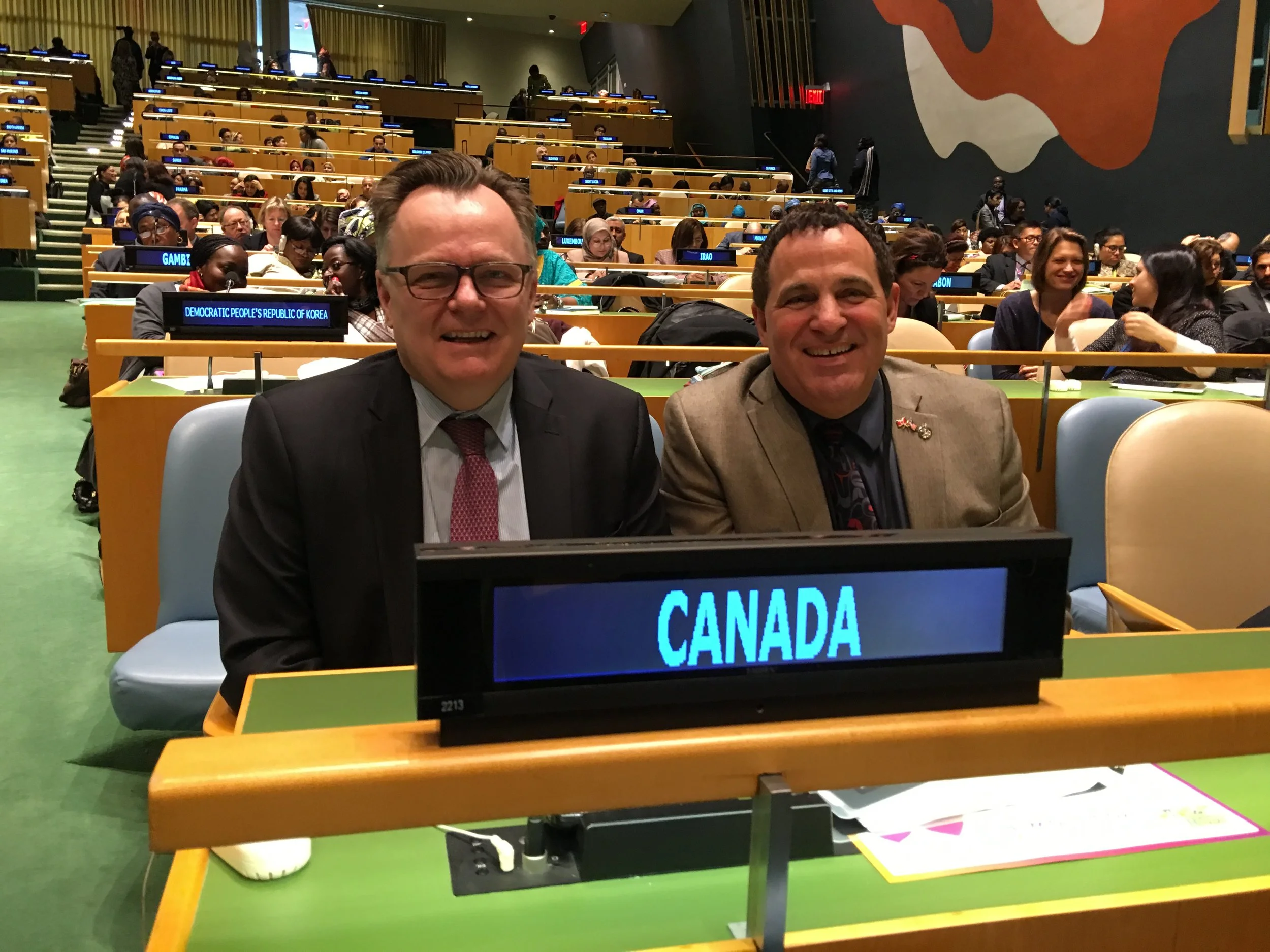Two men in suits sitting at a table with a sign that reads 'Canada' in a large conference room filled with delegates.