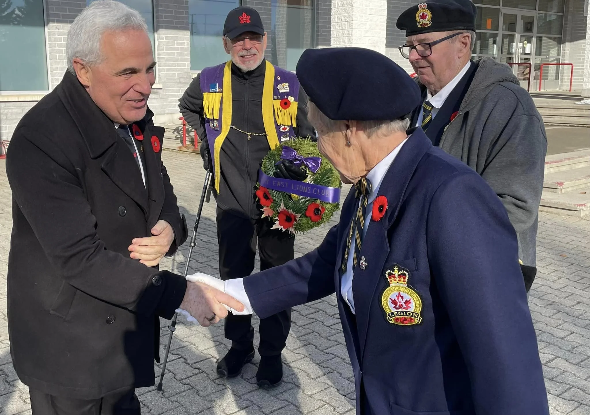 Five men outdoors, dressed in formal and veteran attire, shaking hands and exchanging a wreath with poppy pins, during a remembrance or memorial event.