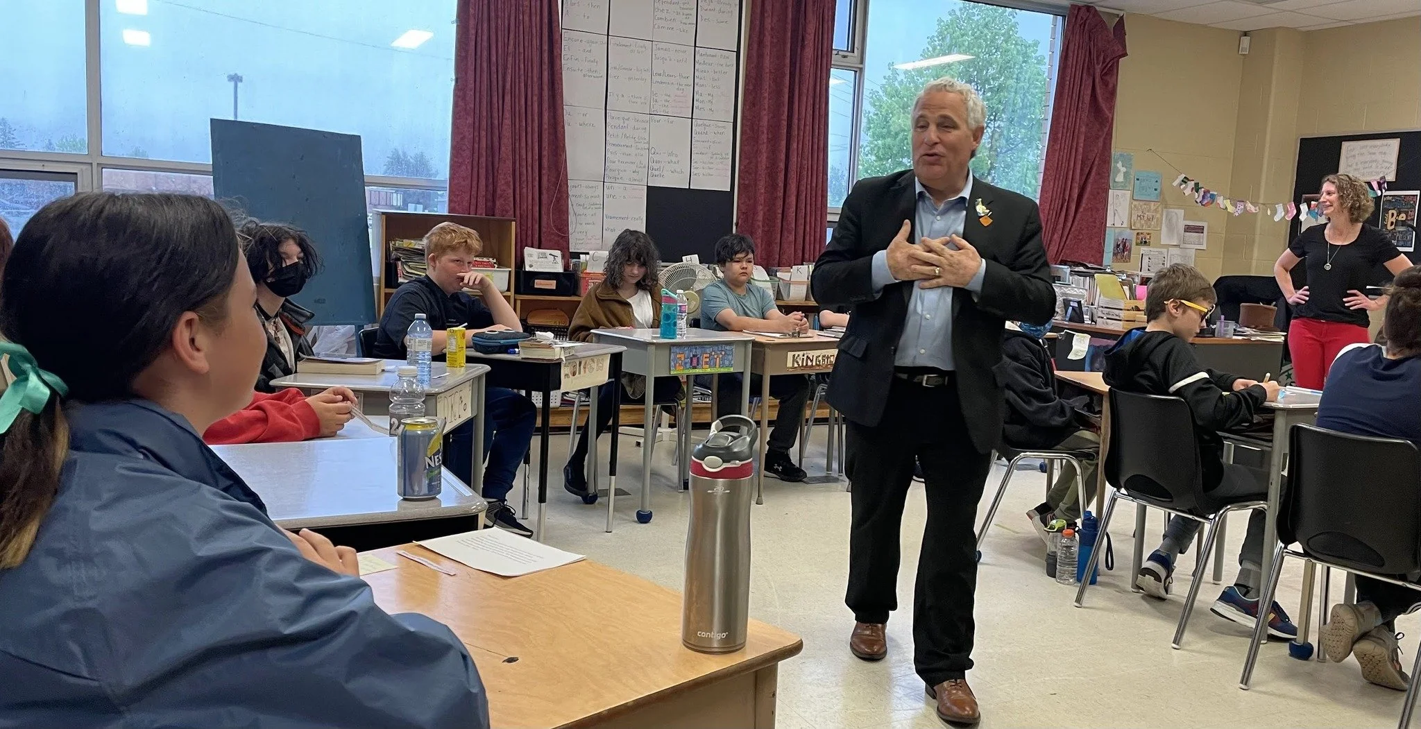 A classroom scene with students sitting at desks, listening to a man in a black suit and a woman in red pants, who are speaking at the front. The classroom has large windows, red curtains, and educational posters on the walls.