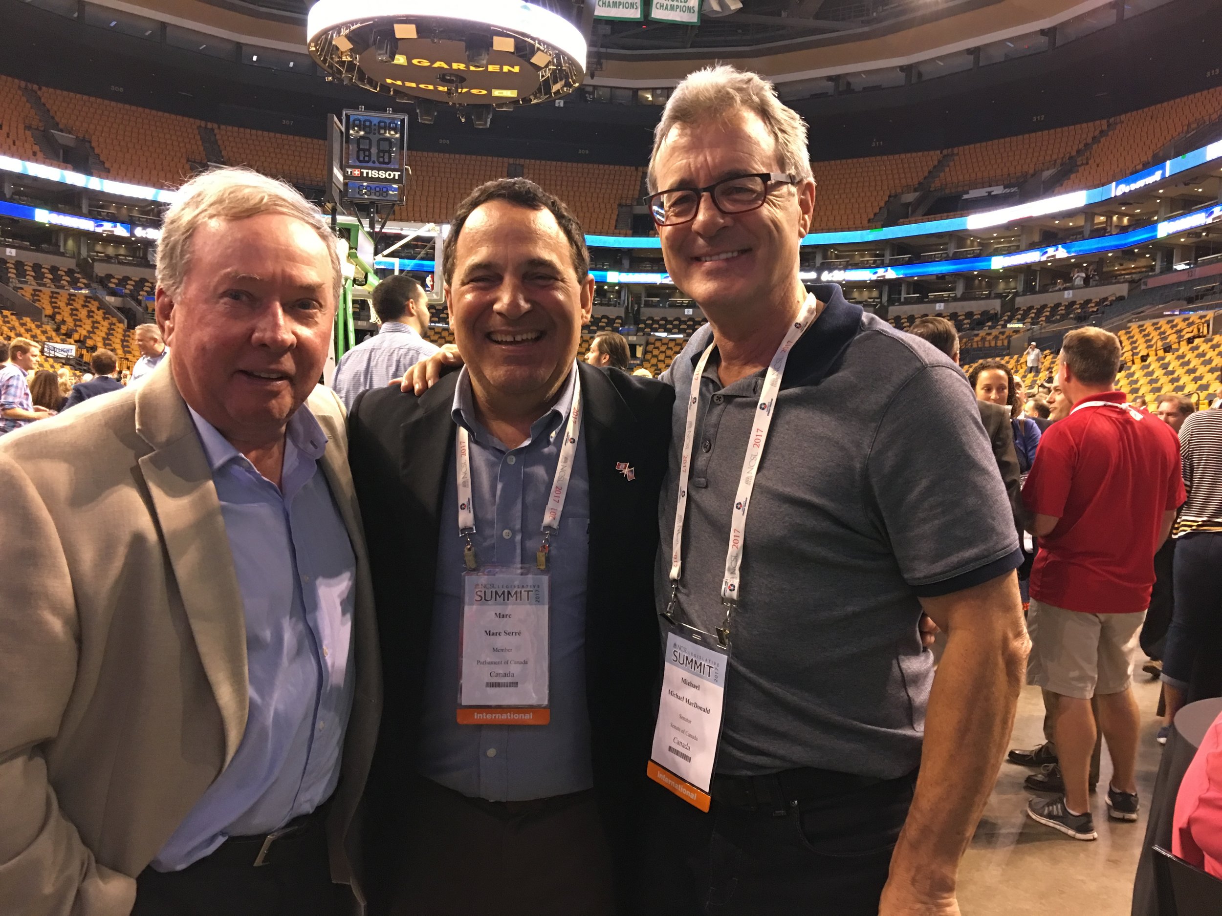 Three men standing together, smiling, inside an arena with yellow seating and a digital scoreboard overhead. The man on the left is wearing a beige blazer, the man in the middle is in a black suit, and the man on the right is wearing a gray polo shir