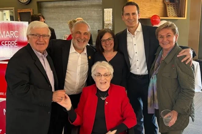 Group of six people, five standing and one sitting, smiling and posing together indoors, with a red sign in the background.