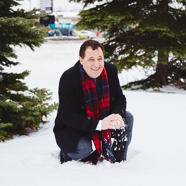 A man in a black coat, red plaid scarf, and gray pants crouching in the snow and holding snow in his hands, smiling.