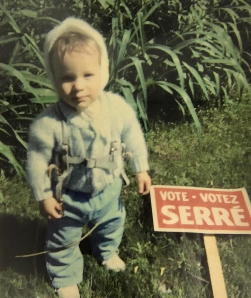 A young child wearing a knit hat and a cozy sweater stands outdoors on grass next to a campaign sign that reads 'VOTEZ SERRE,' with tall grass or crops in the background.