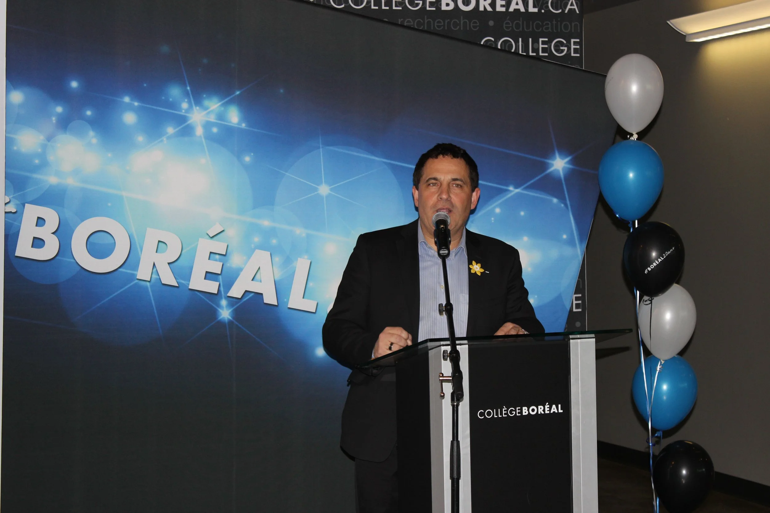 Man in a black suit speaking at a podium with a blue background that says 'Collège BOREAL' and a cluster of black, white, blue, and gray balloons to the right.