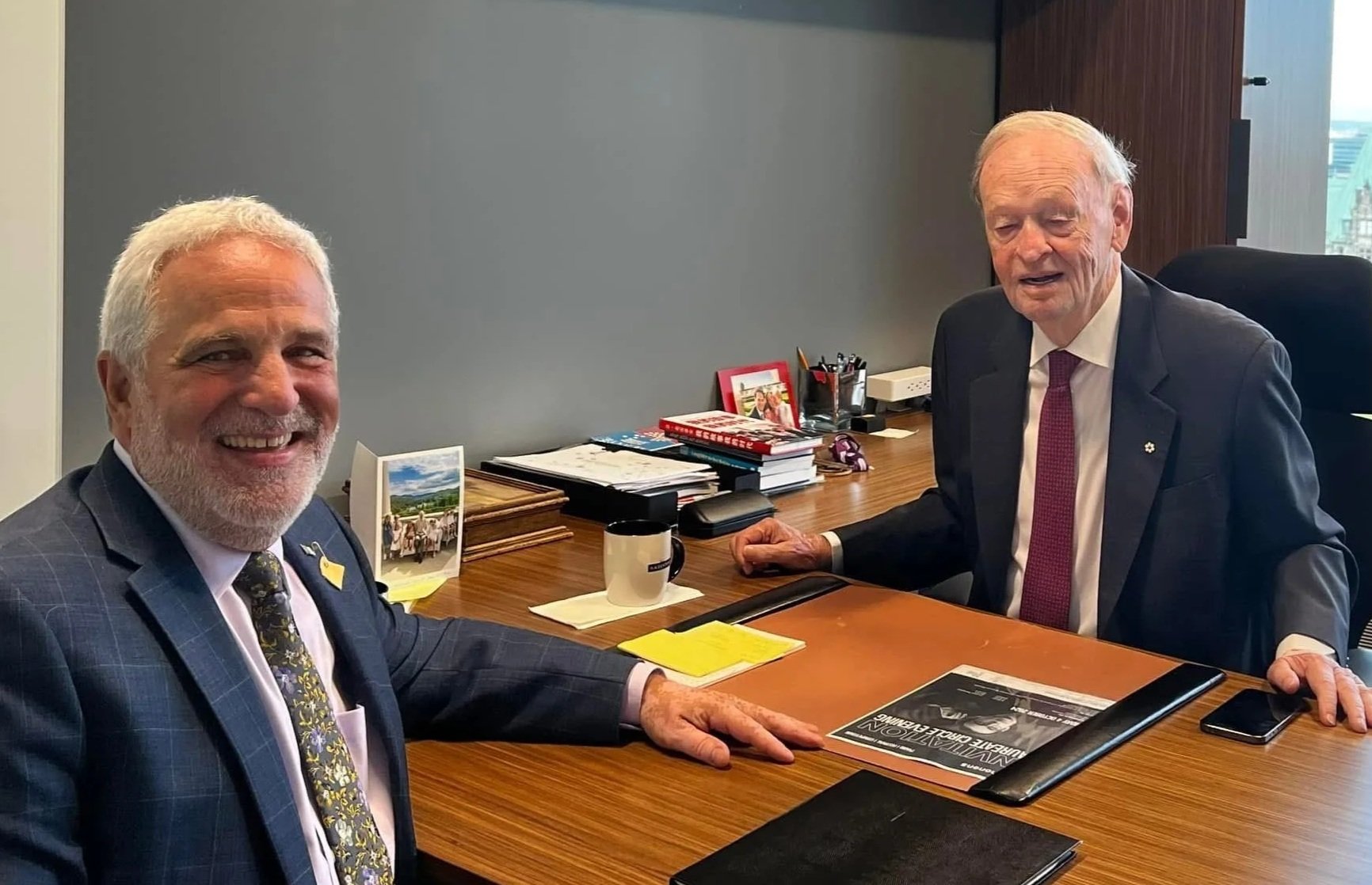Two older men in suits sitting at a wooden office desk, smiling, with documents, books, a coffee mug, and electronic devices on the desk.