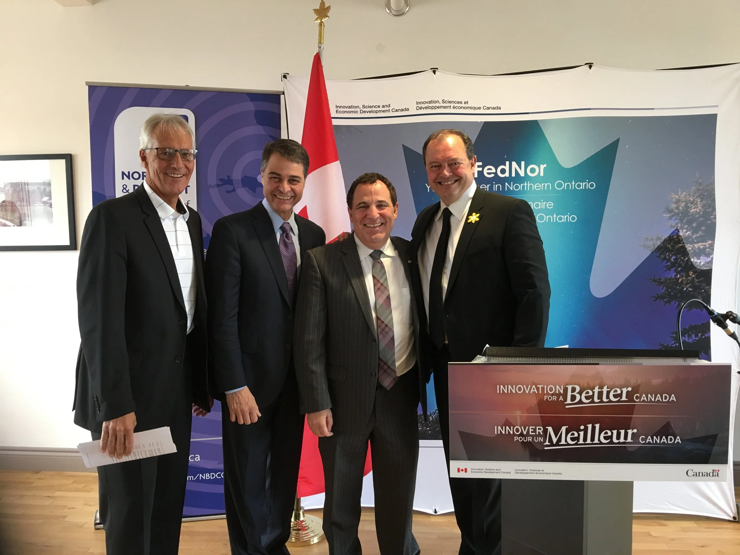 Four men standing in front of a Canadian flag and banners, dressed in suits, smiling for the camera at a formal event.
