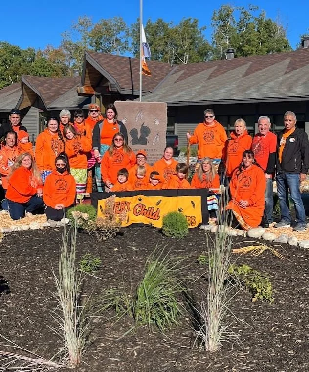 Group of children and adults wearing matching orange sweatshirts posing outdoors in front of a stone monument. They are holding a yellow banner that reads 'Every Child Matters.' The setting includes a landscaped area with plants and rocks, and a buil