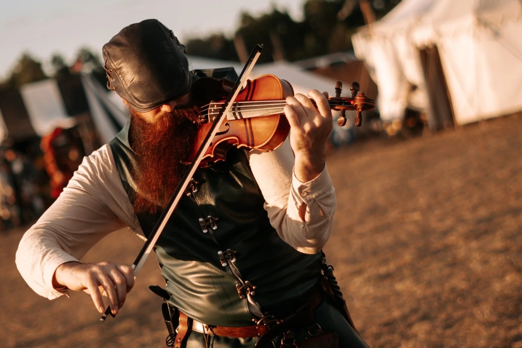A man with a red beard playing the violin outdoors, wearing a black hat, a vest, and a long sleeve shirt, with tents in the background.