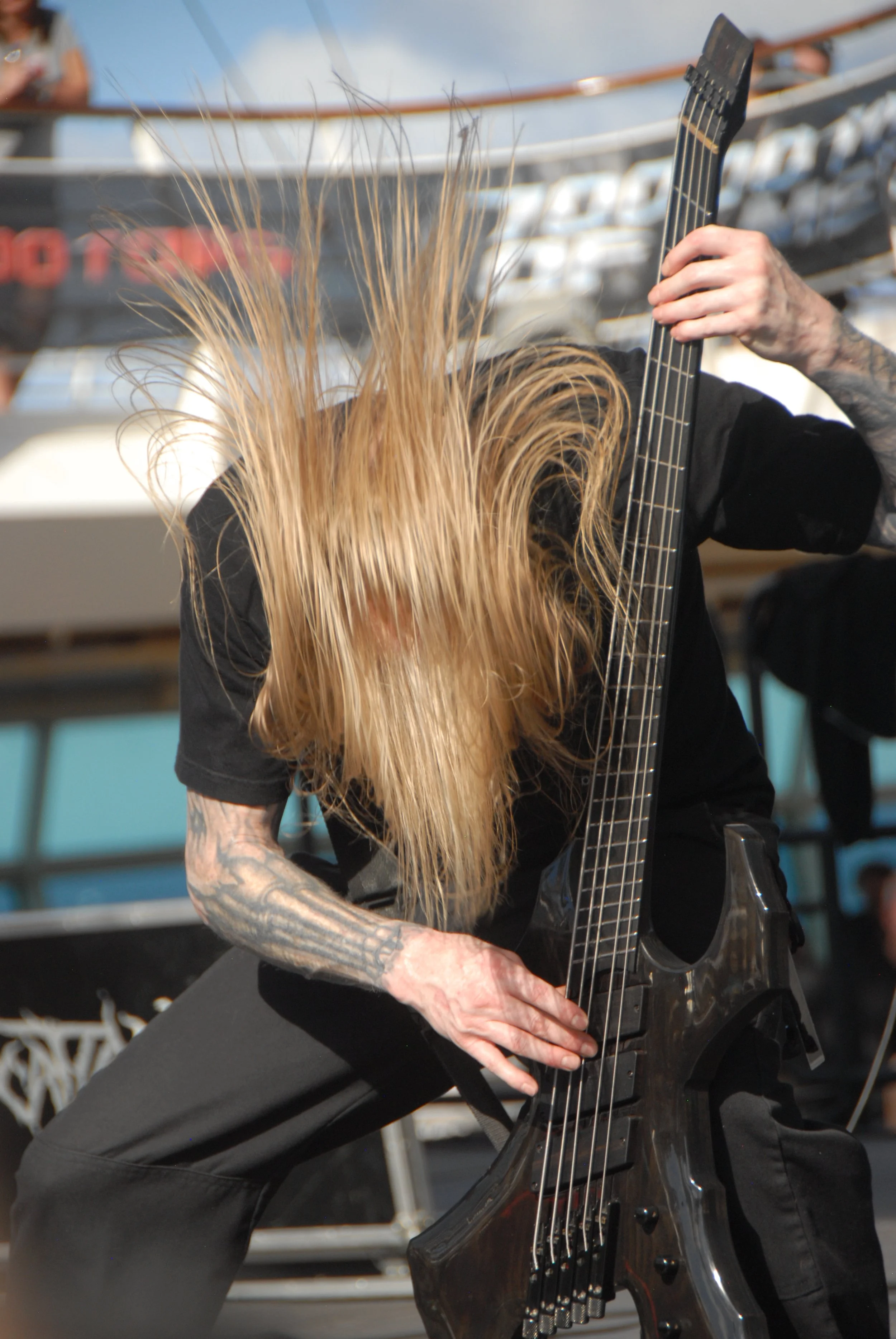 A musician with long blonde hair, black tattoos on their arm, wearing a black shirt, playing an electric bass guitar during a live performance outdoors.