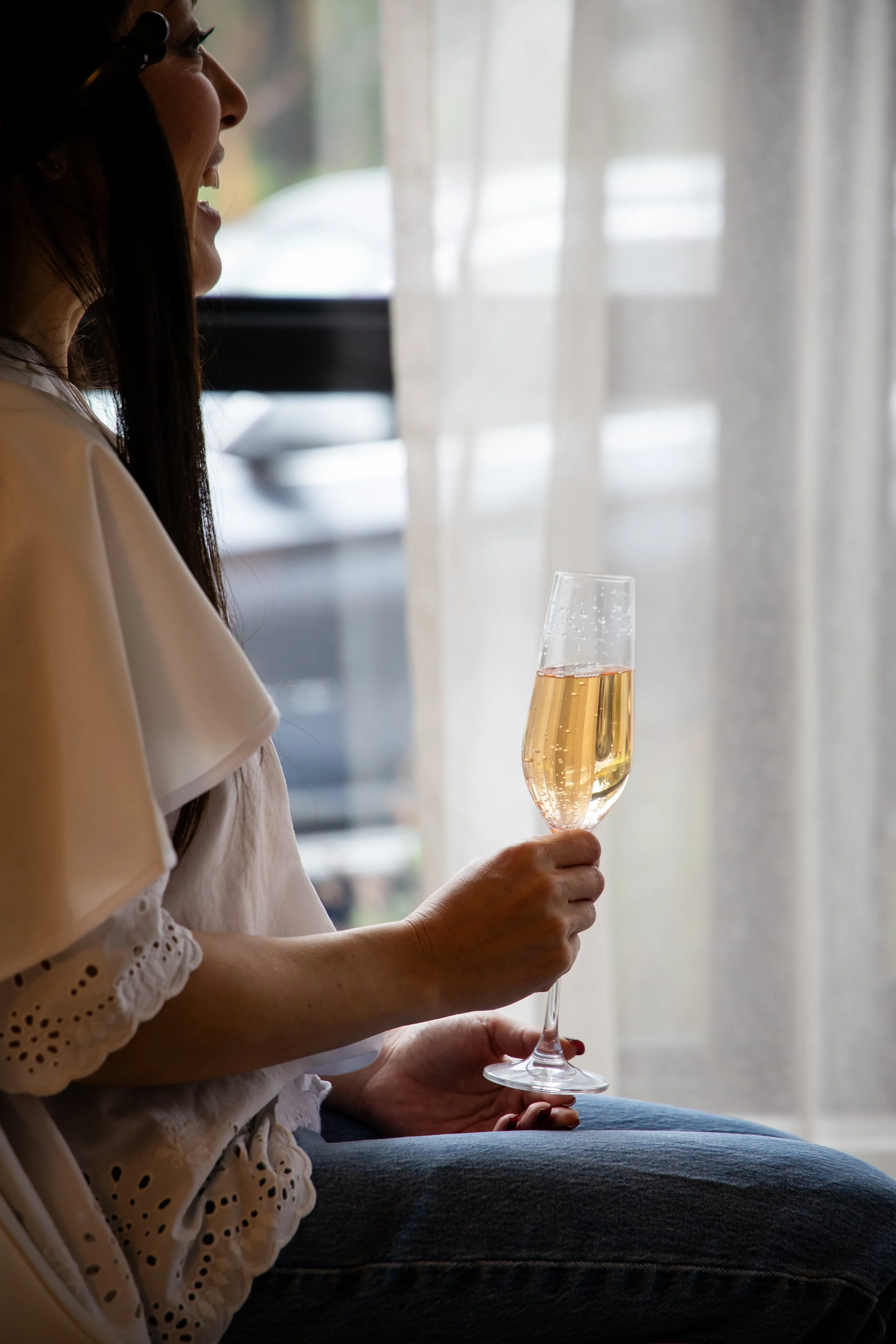 A woman sitting indoors near a window with sheer curtains, holding a glass of champagne.