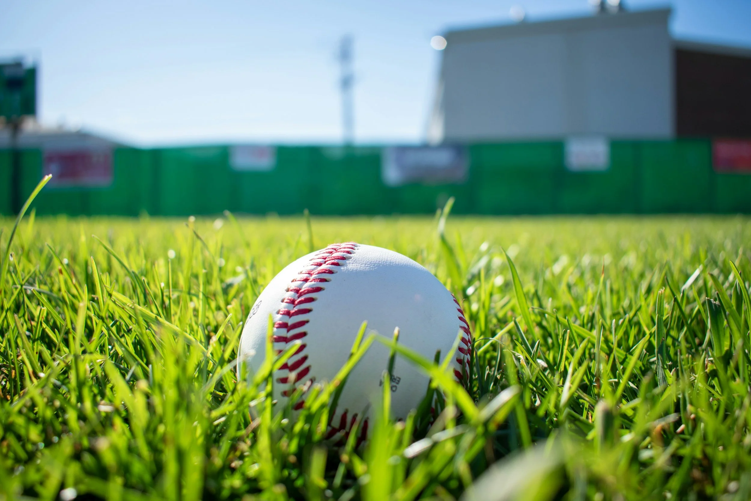 Close-up of a baseball on green grass with a fence, building, and blue sky in the background.