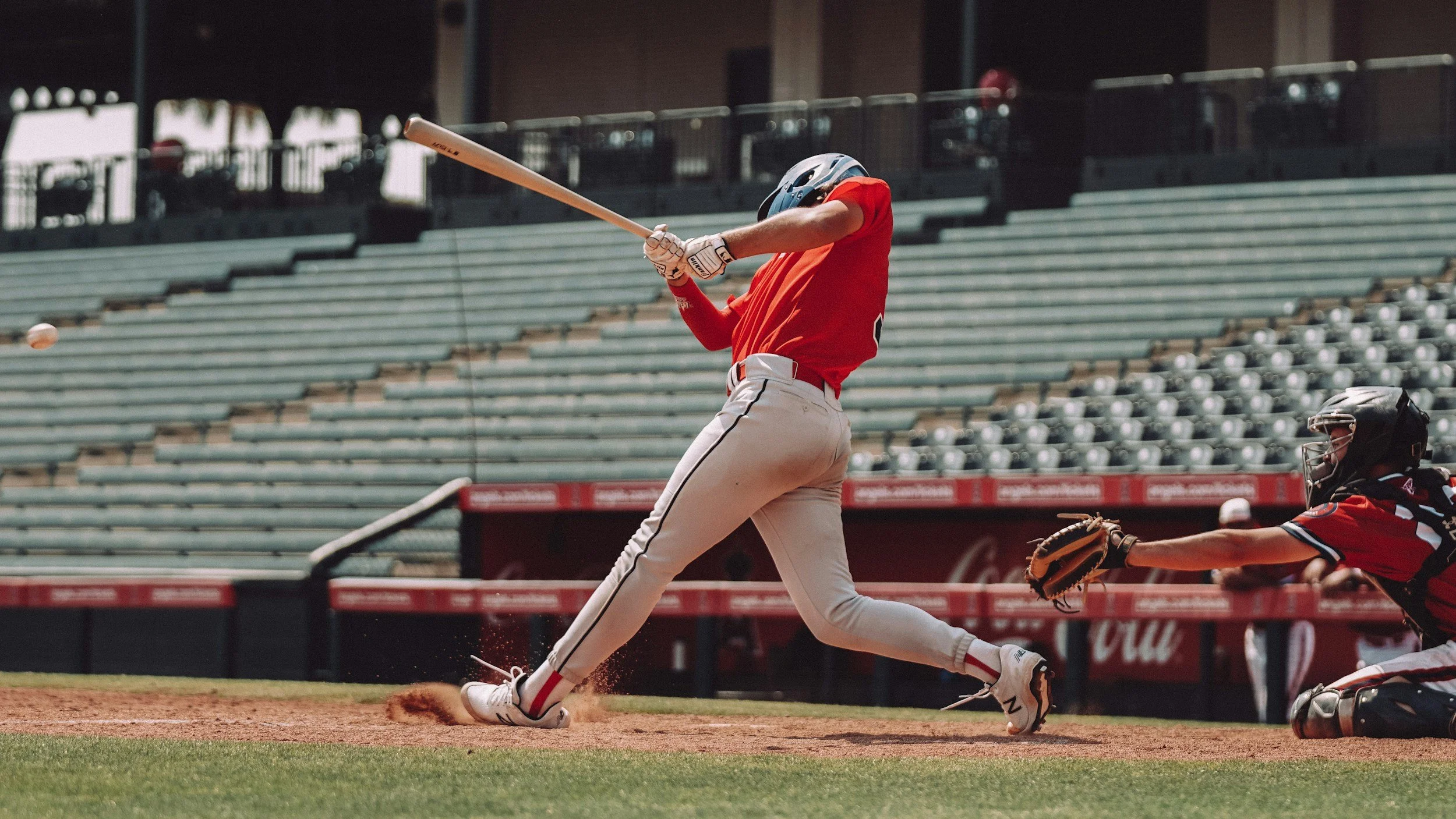A baseball player in a red uniform hits the ball with a bat during a game, while a catcher in red and black gear reaches out to catch the ball on a baseball field with empty stands in the background.