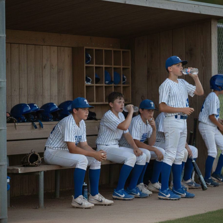 Baseball players sitting on a team bench, wearing striped uniforms and blue helmets, with some drinking water and holding bats, under a wooden sheltered area.