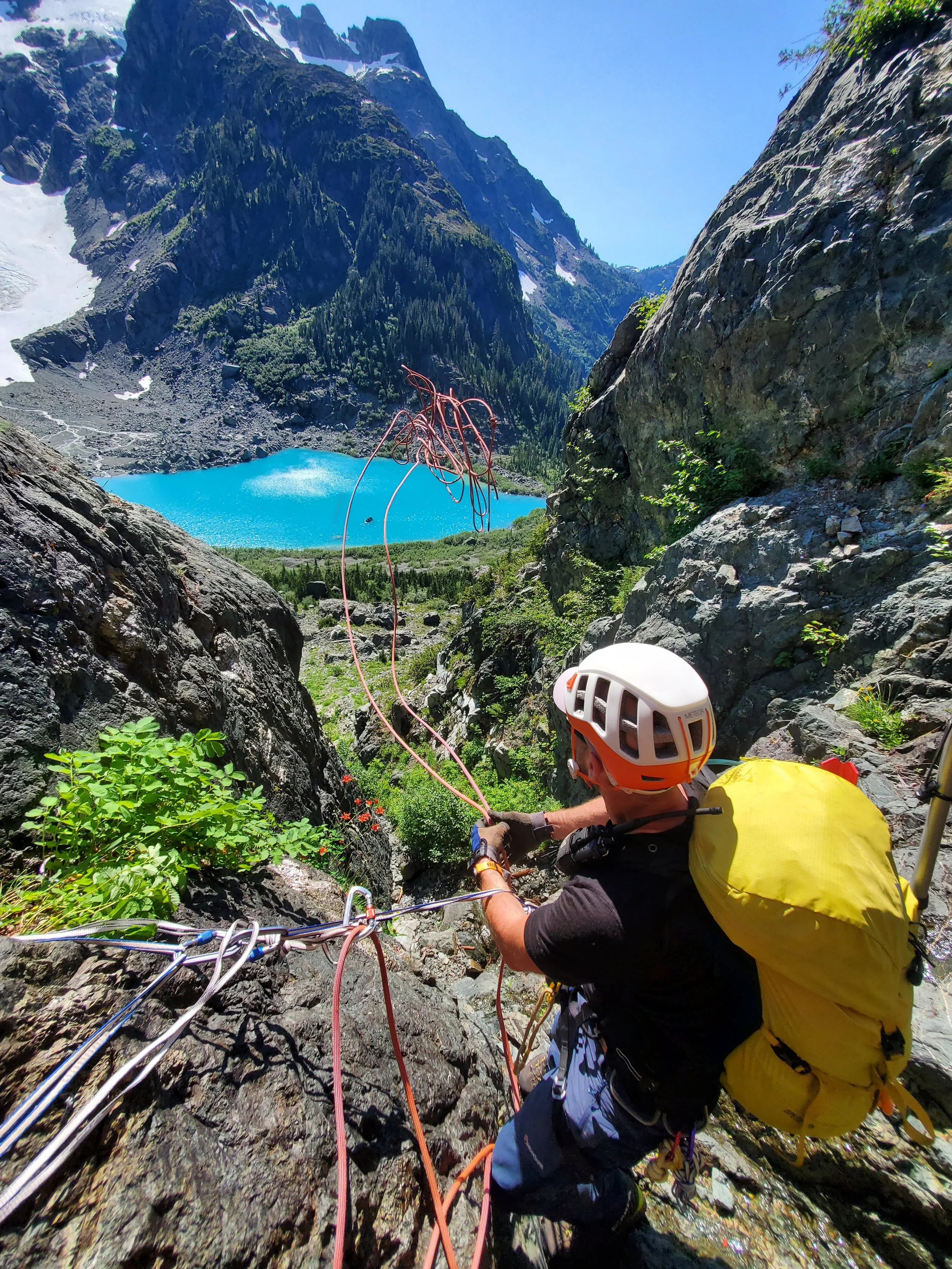 A climber with a white helmet and yellow pack ascending a rocky mountain, overlooking a turquoise lake surrounded by green trees and snow-capped peaks.