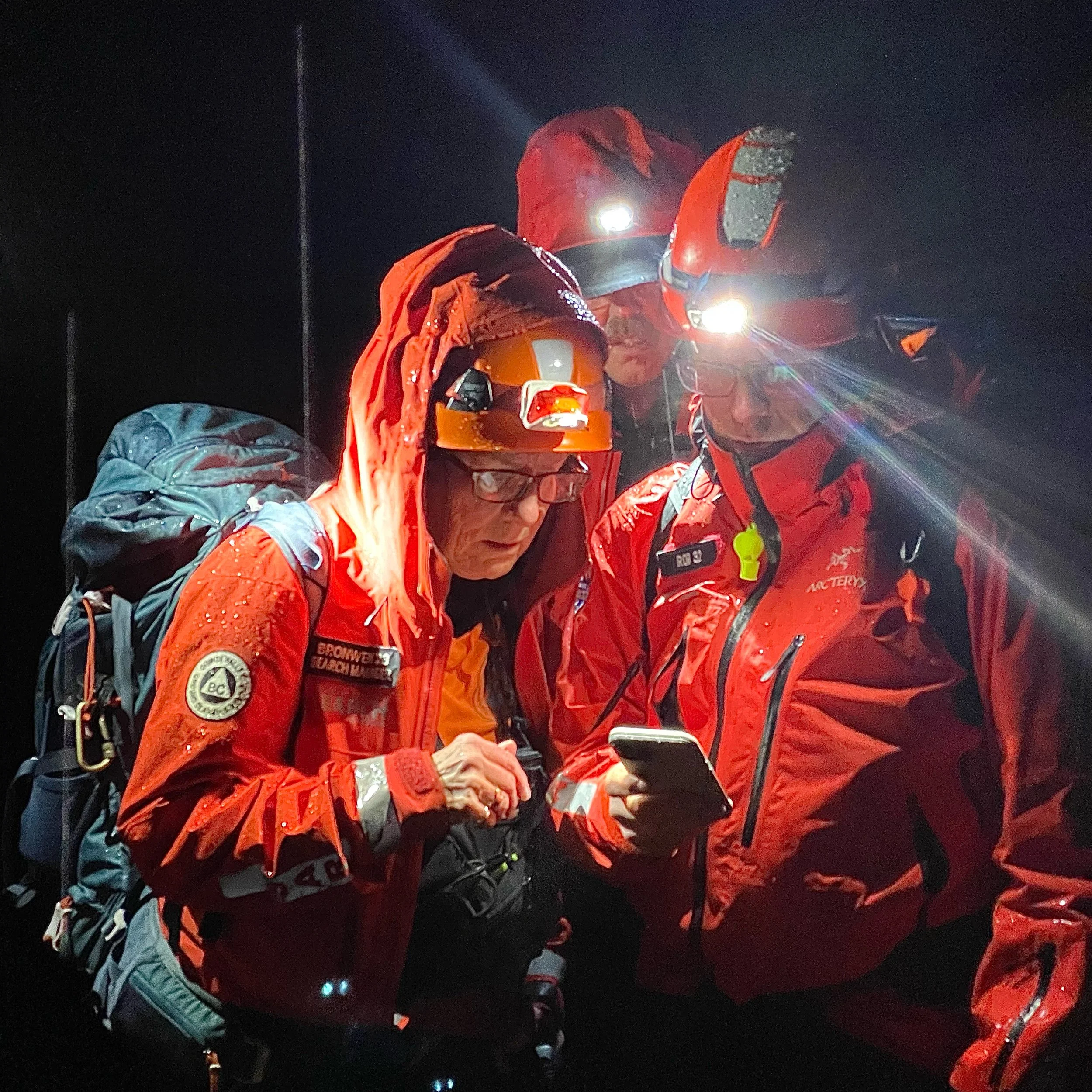 Three rescue workers in red jackets with headlamps and helmets, one with glasses, looking at a smartphone at night, wearing backpacks and gear.