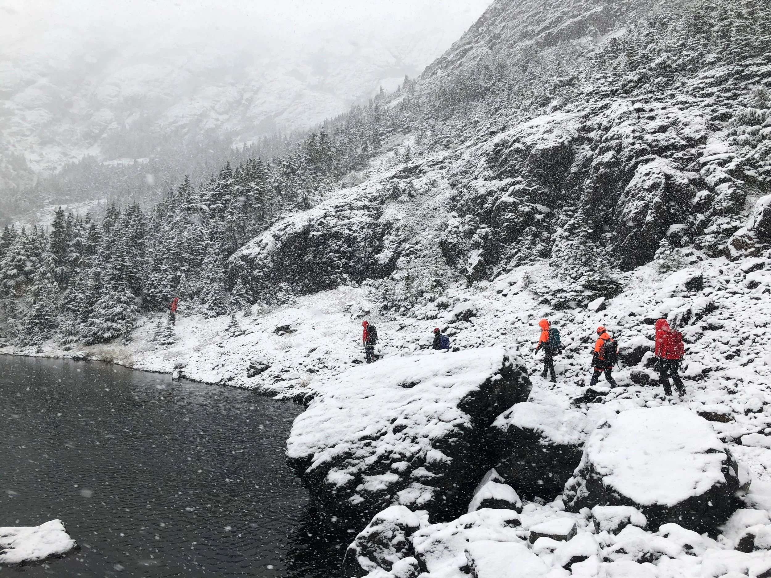 A group of hikers wearing orange jackets trekking along a snowy rocky shoreline by a lake in a mountainous, snow-covered landscape.