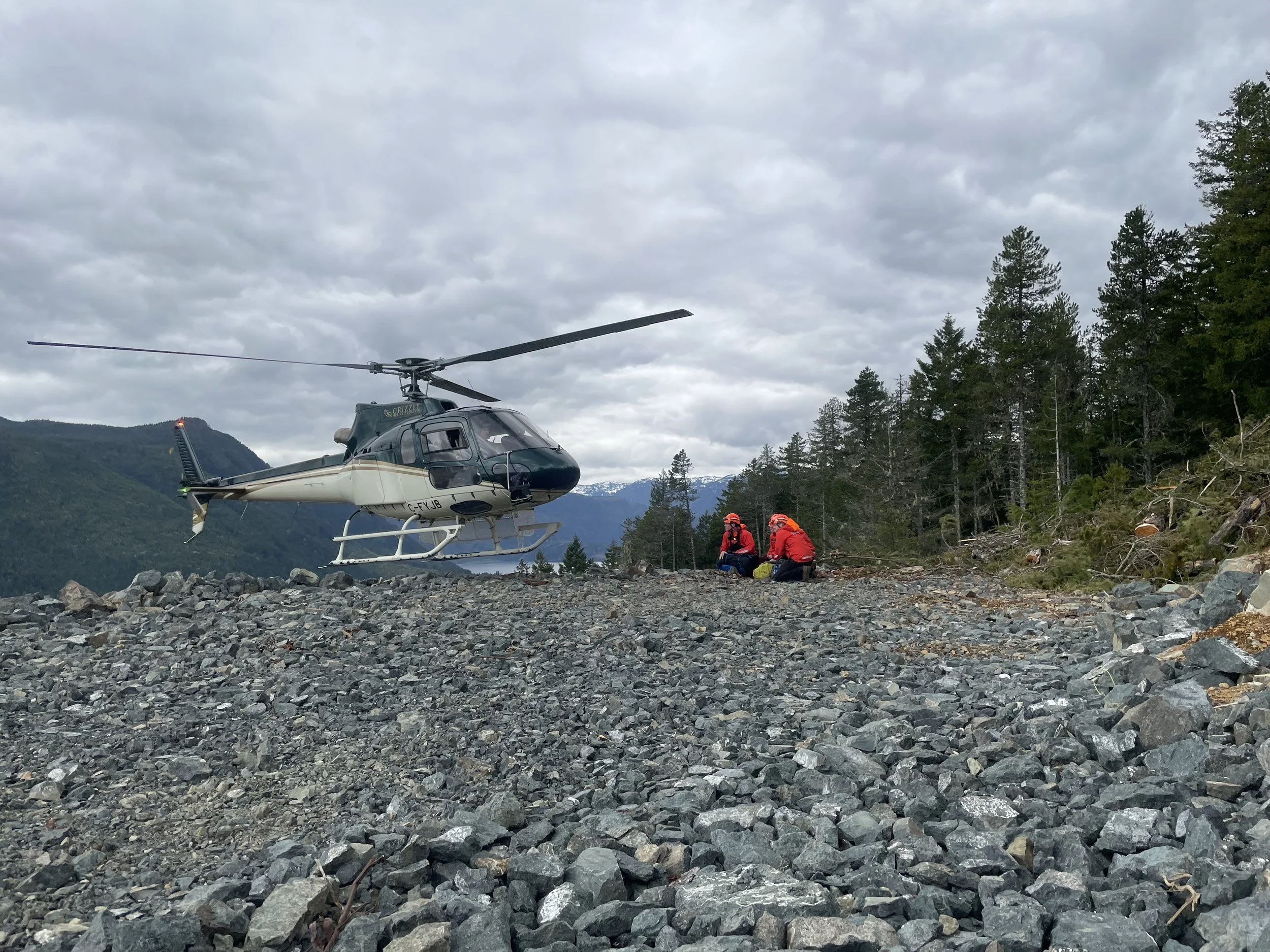 A helicopter flying over rocky terrain with two rescue workers in red jackets and helmets sitting and filling out paperwork, surrounded by trees and mountains under a cloudy sky.