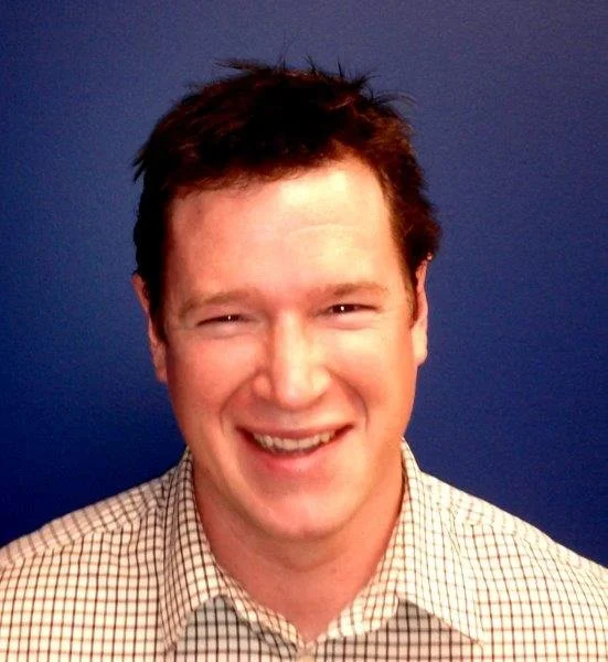 A smiling man with short, dark hair, wearing a checkered shirt, standing against a blue background.