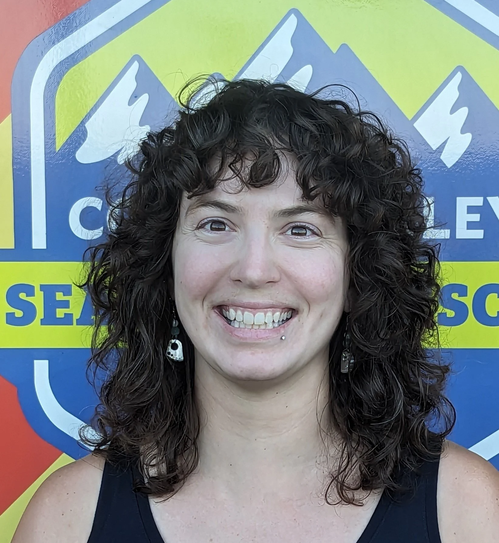 Smiling woman with short hair and a scarf, standing outdoors in front of a painted brick wall.