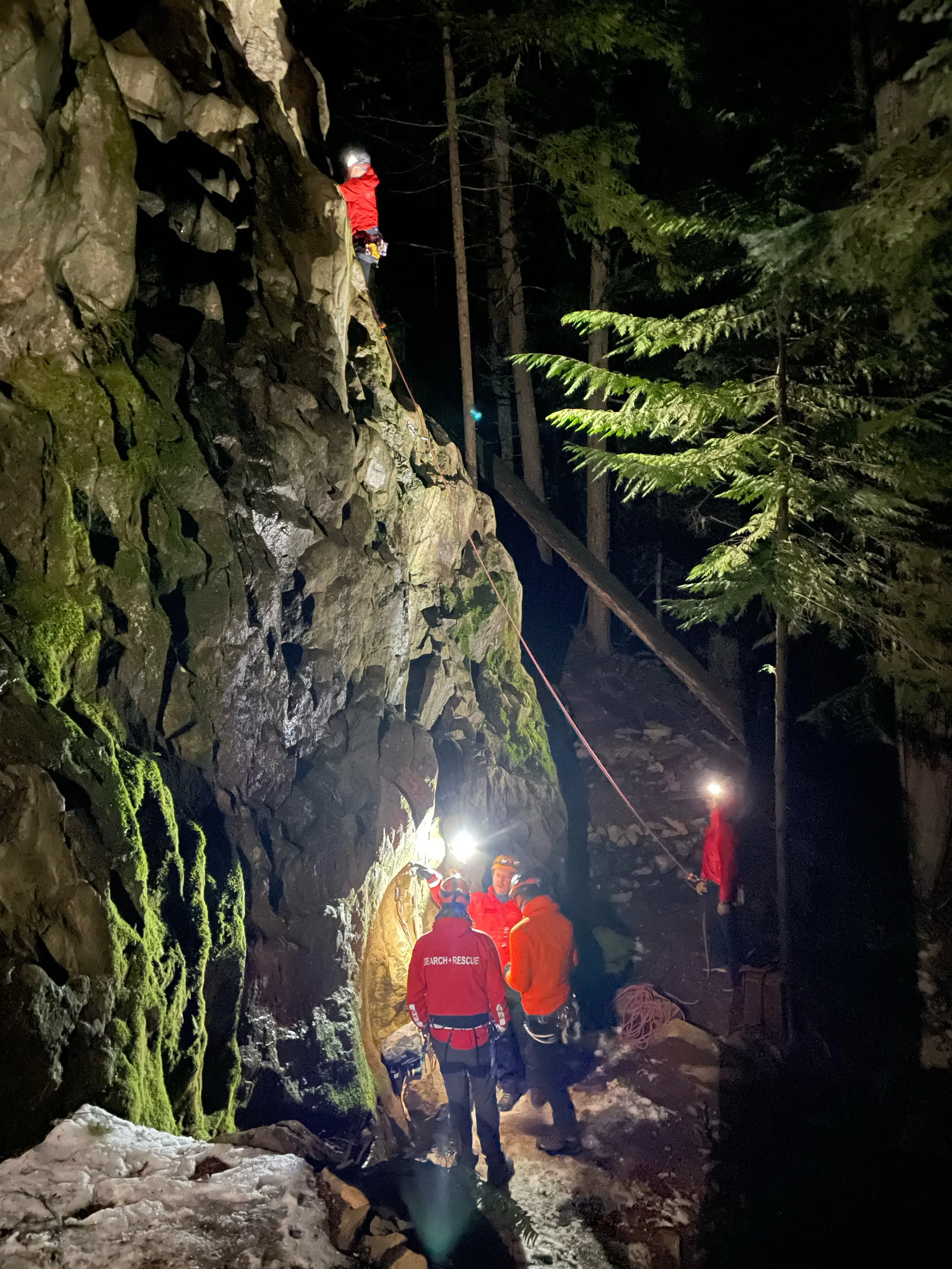 Night scene of a cliff rescue operation with rescue workers in helmets and headlamps near a moss-covered rocky cliff, surrounded by tall trees.