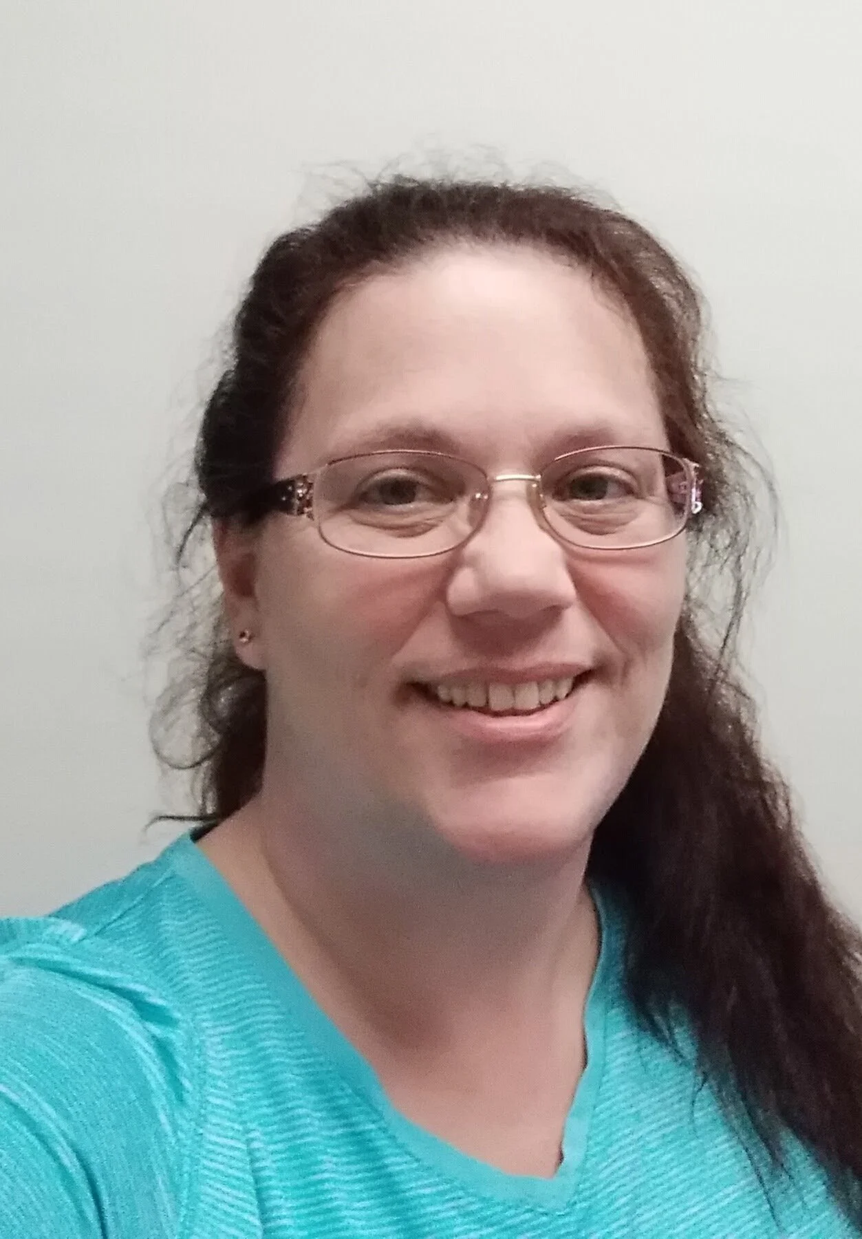 A woman with glasses, long brown hair, and a teal shirt smiling at the camera against a plain wall.