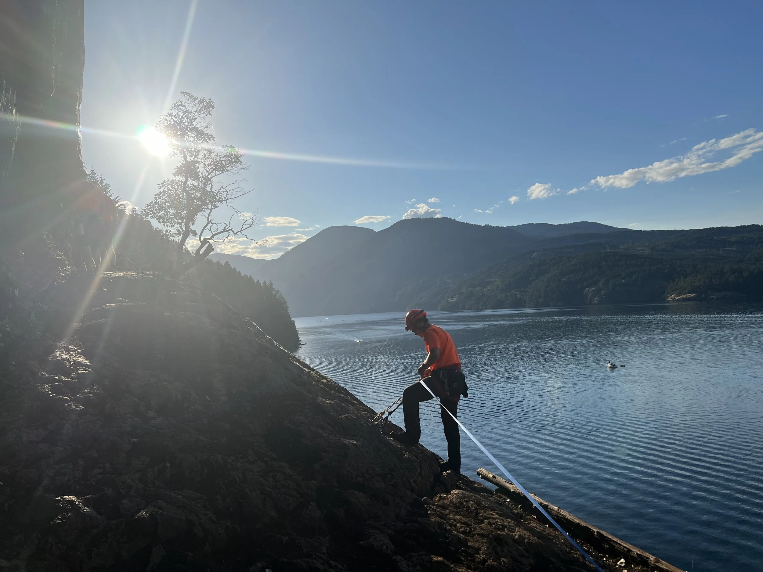 A person in a red shirt and helmet is climbing a rocky slope near a lake with mountains in the background under a blue sky.