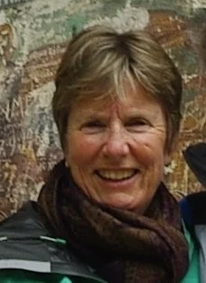 Smiling woman with short hair and a scarf, standing outdoors in front of a painted brick wall.
