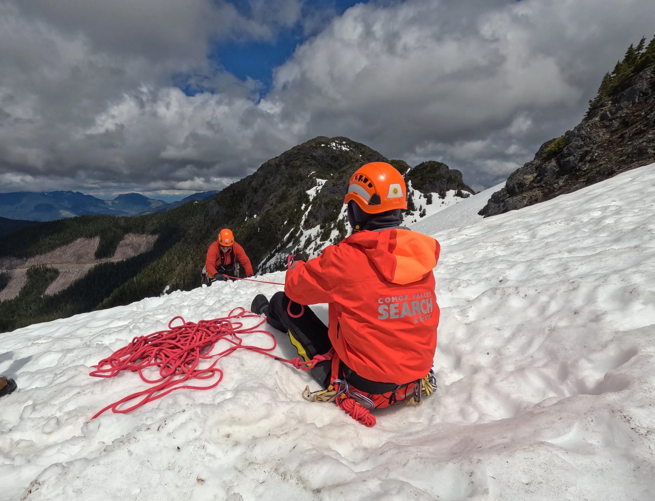Two rescue team members in orange jackets and helmets on a snowy mountain, one sitting and the other climbing, with ropes and mountain scenery in the background.