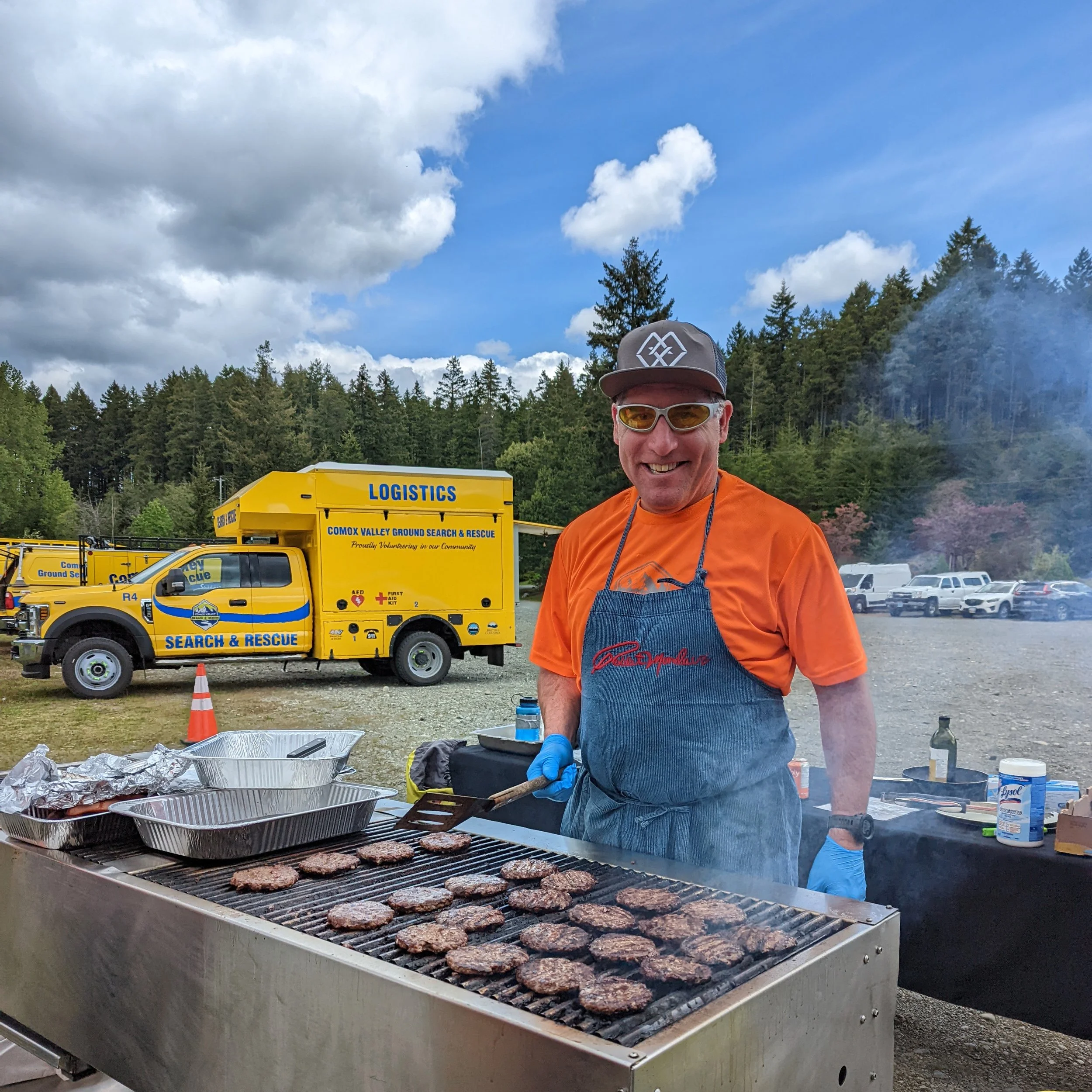 A man grilling hamburgers at an outdoor event with a yellow search and rescue vehicle in the background and a parking lot with cars, trees, and a cloudy sky.