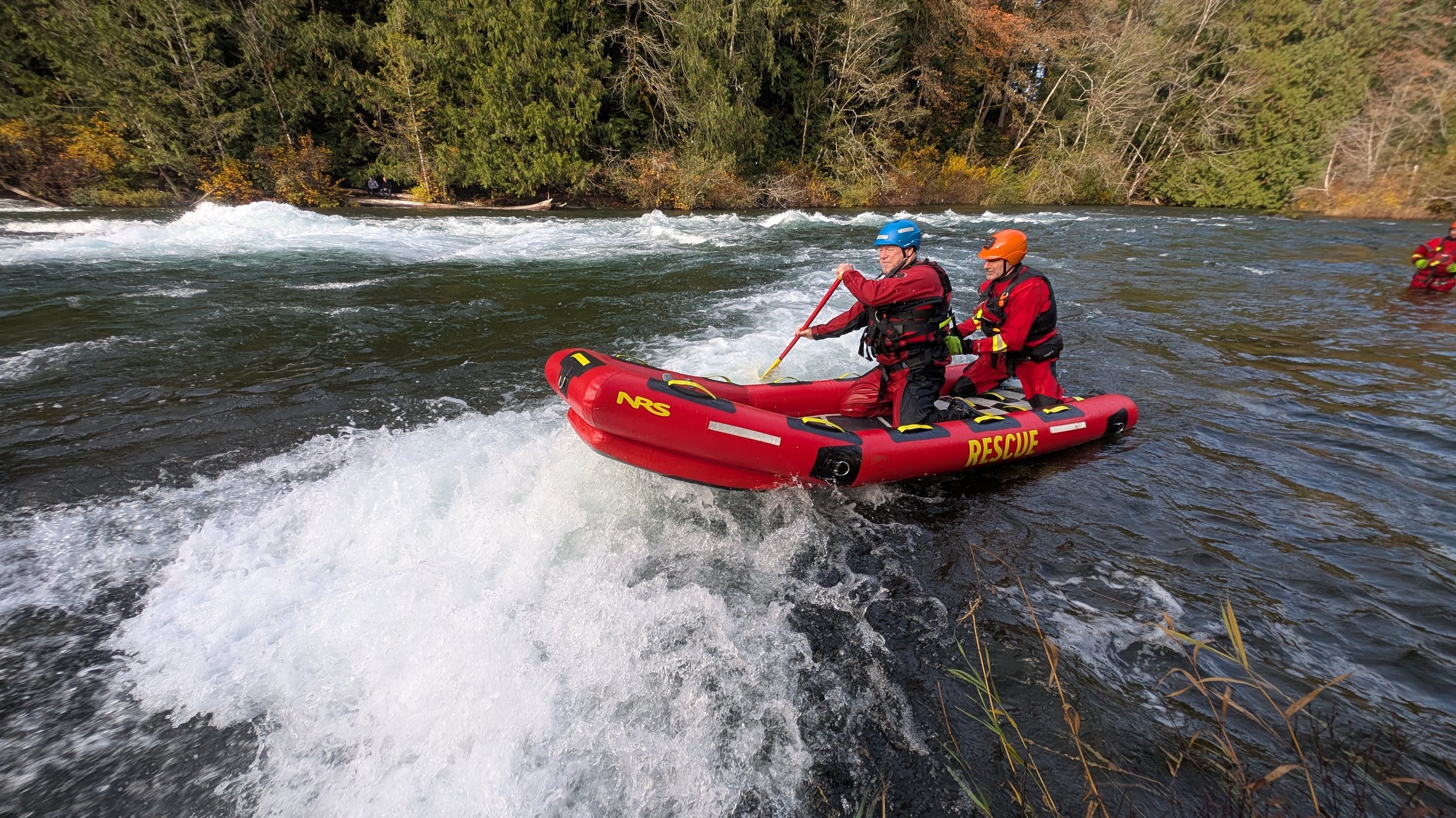 Two rescue workers in red uniforms with helmets riding a red rescue boat on a river with trees in the background.