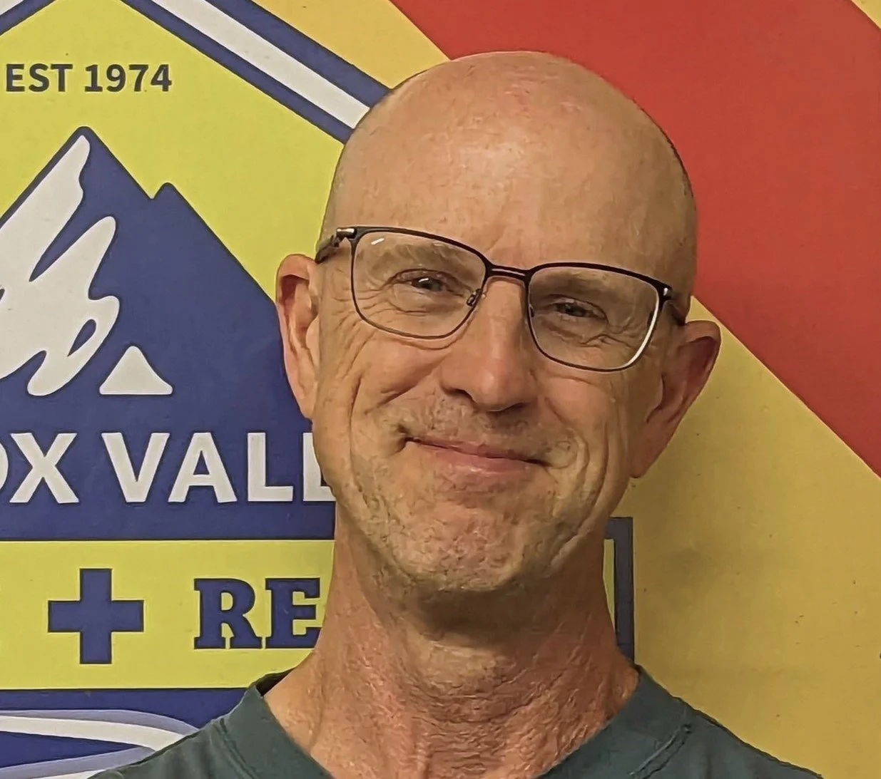 A smiling elderly man with glasses standing in front of a colorful sign that reads "Snow Valley" and includes a mountain graphic, the text "EST 1974," and additional text and symbols.