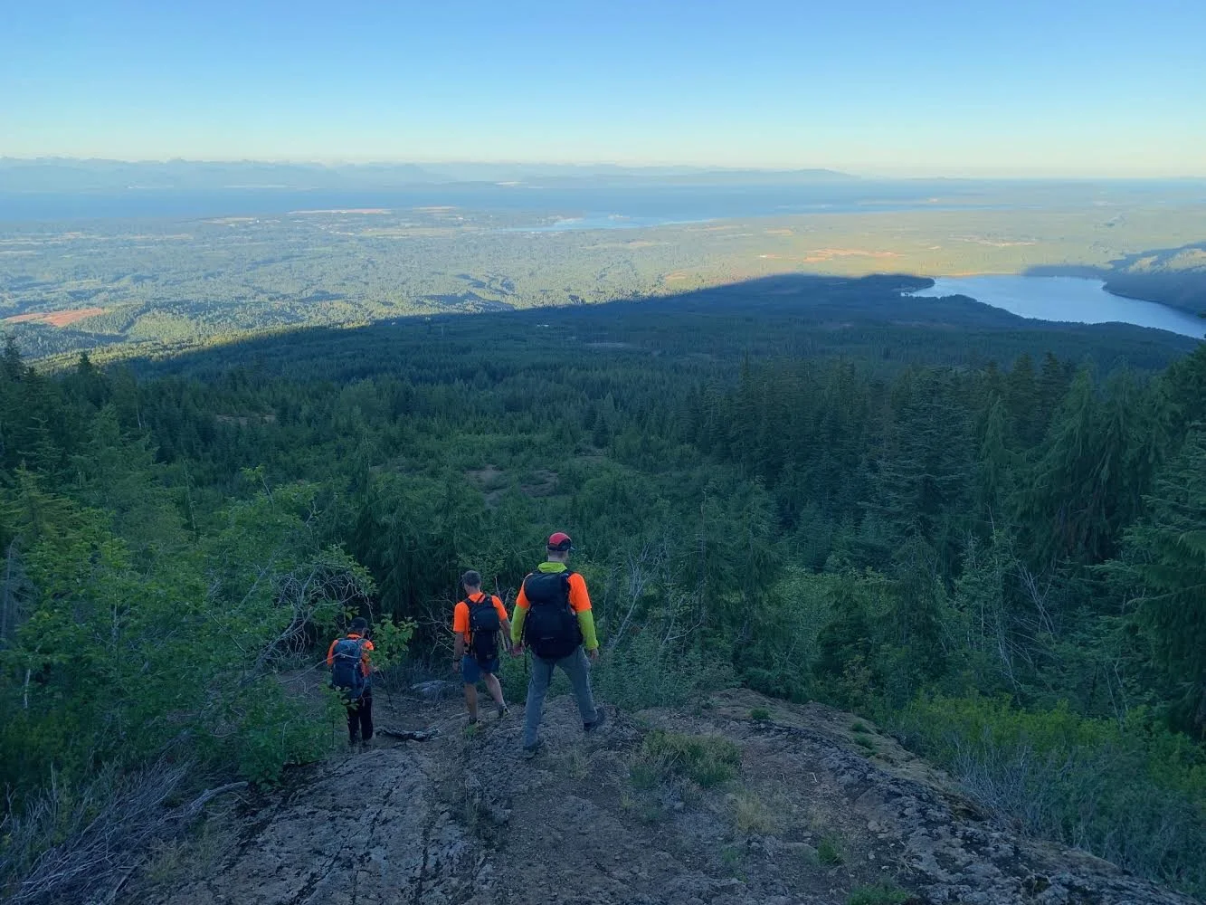 Four hikers with backpacks descend a rocky trail through a dense green forest, overlooking a wide valley with rivers, lakes, and distant mountains under a clear blue sky.
