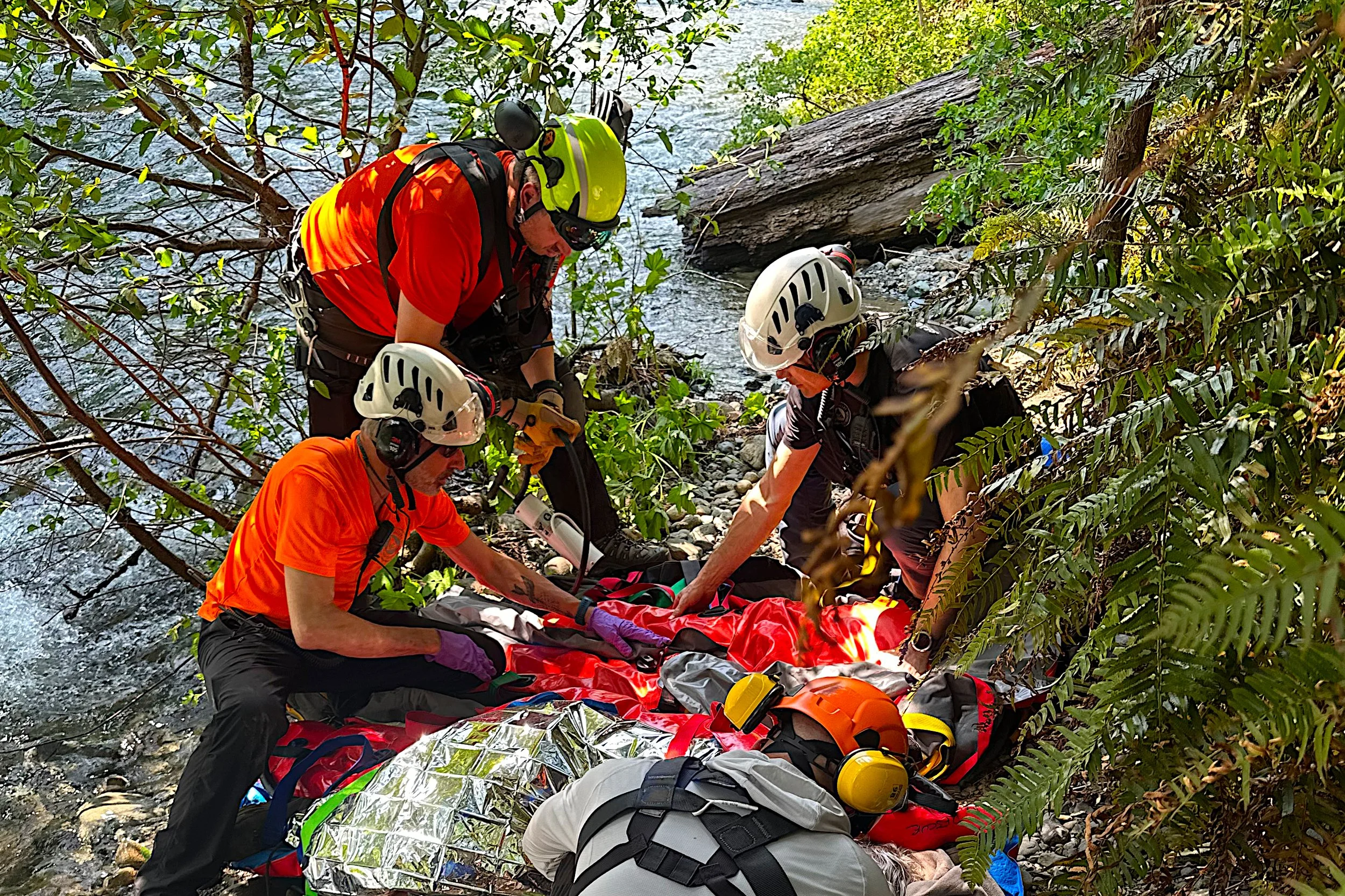 Rescue team members in helmets and gear attending to an injured person on the ground by a river in a wooded area.