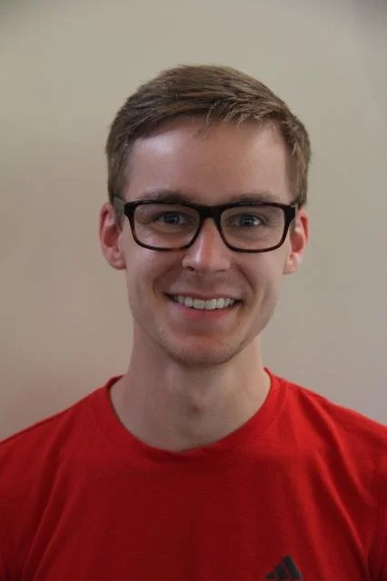 Young man wearing glasses and a red shirt smiling at the camera against a plain wall background.