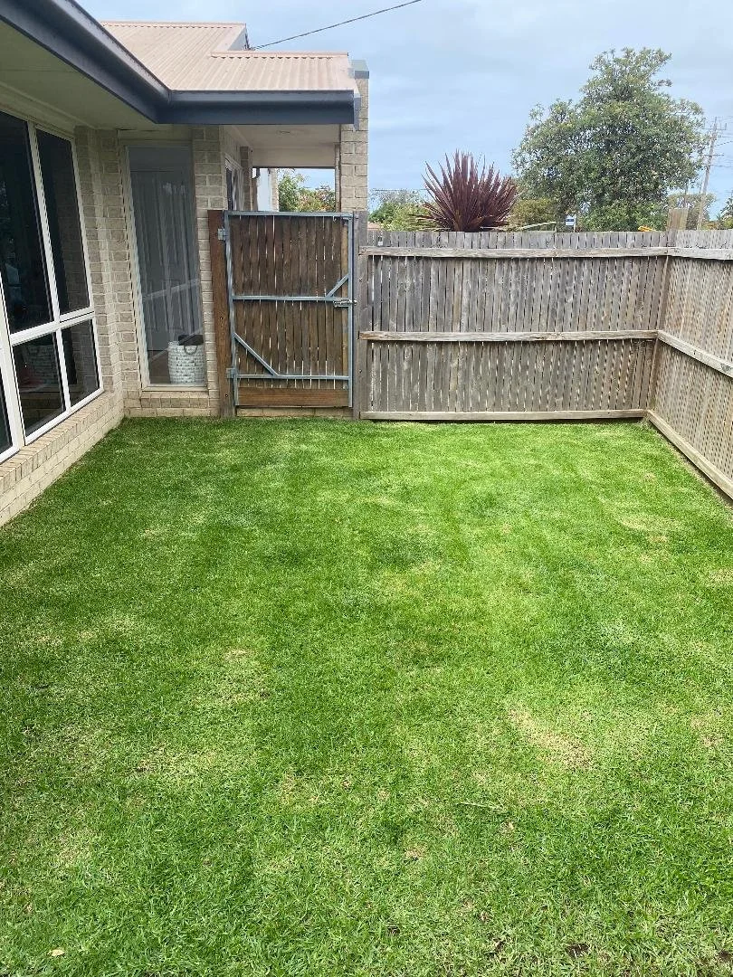 A small backyard with a freshly mowed green lawn, enclosed by a wooden fence, with a sliding glass door and windows on the left side of a brick house, and a metal gate leading outside. There are some trees and a bush in the background.