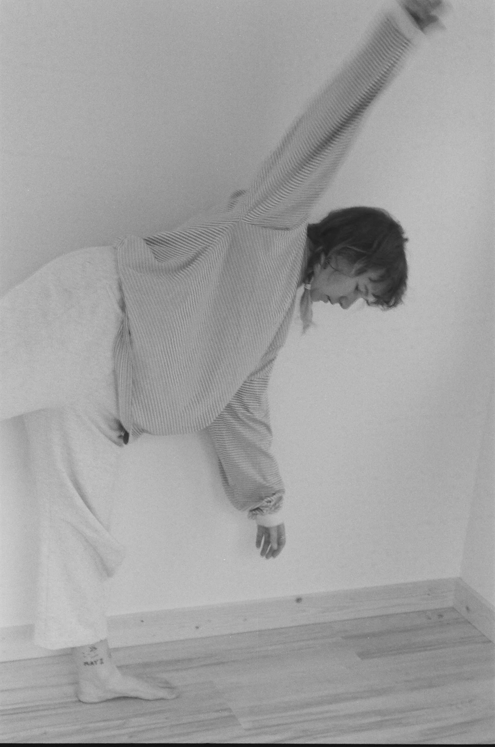 Person wearing pajamas, leaning forward and reaching down, standing on one foot, in a corner of a room with wooden floor and plain wall.