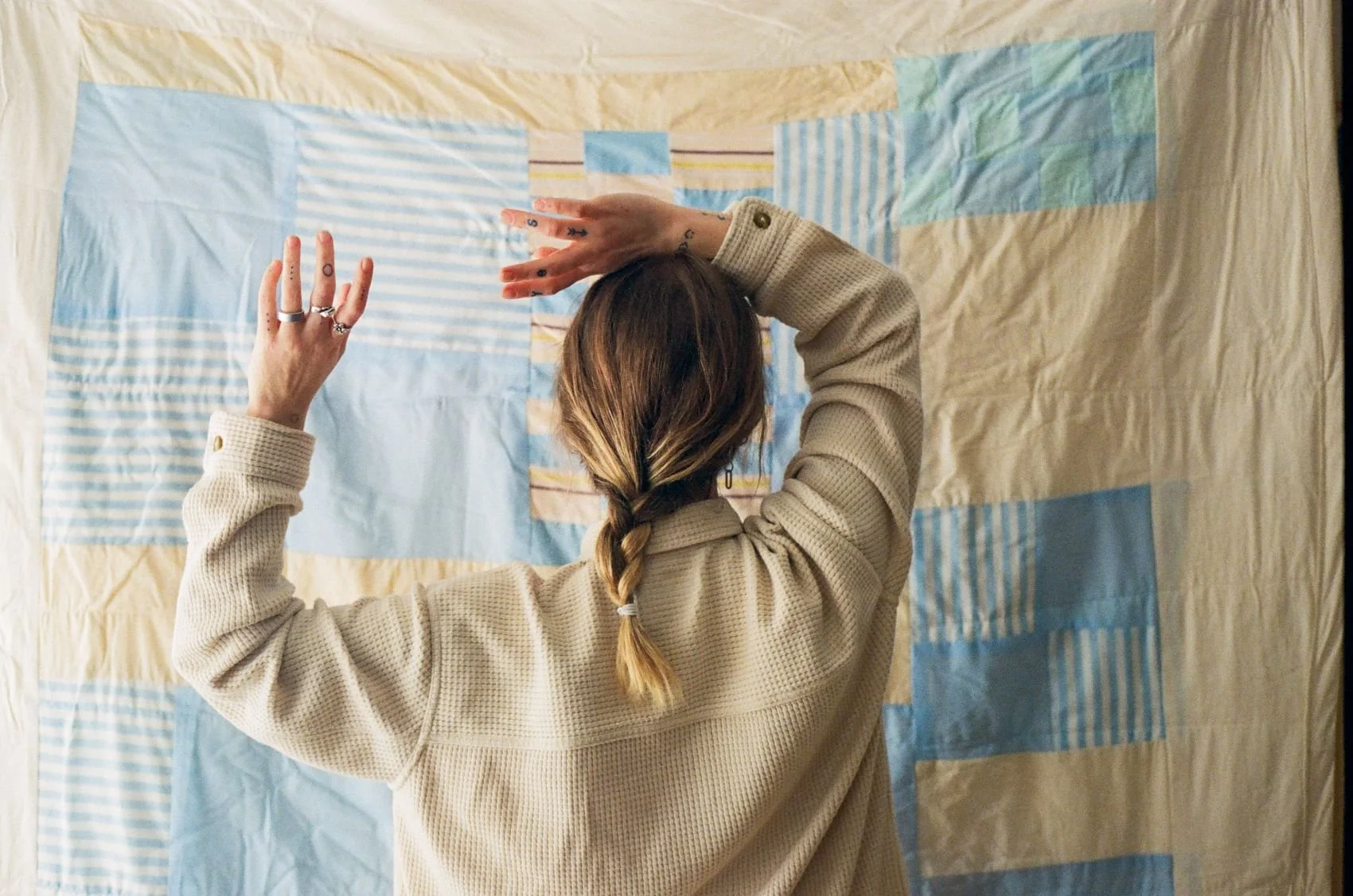 A person with long hair in a braid, wearing a beige sweater, is seen from behind arranging a patchwork quilt or fabric collage on a surface.