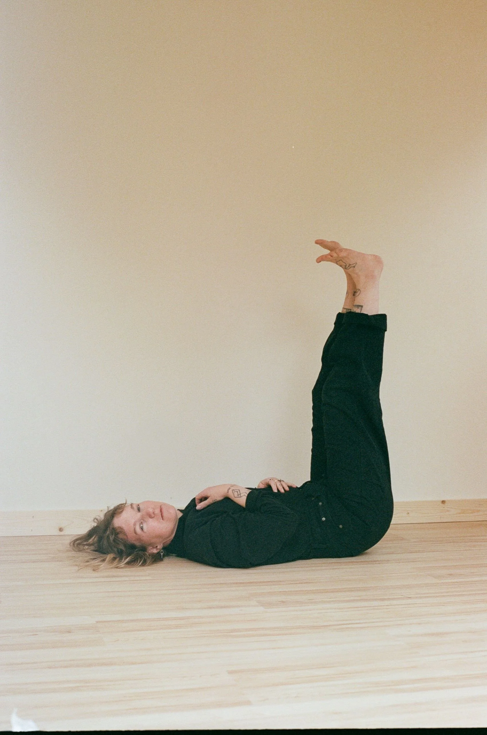 A person with blonde hair lying on their back on a wooden floor, with their legs raised vertically against a plain beige wall, wearing a black outfit.