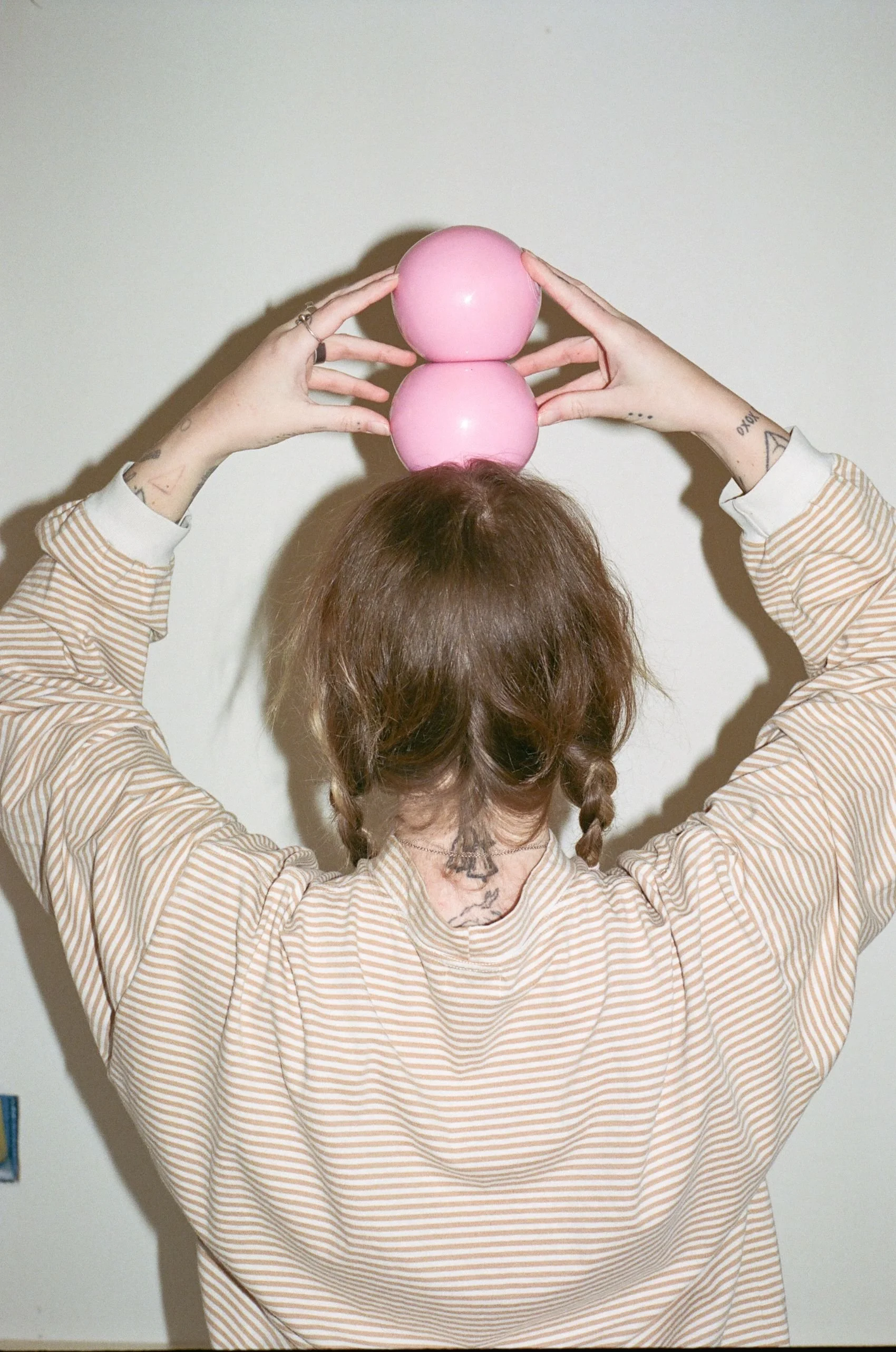 Person with hair in braided pigtails balancing two pink juggling balls on their head, facing away from the camera, wearing a striped shirt, with tattoos visible on their neck and arms.