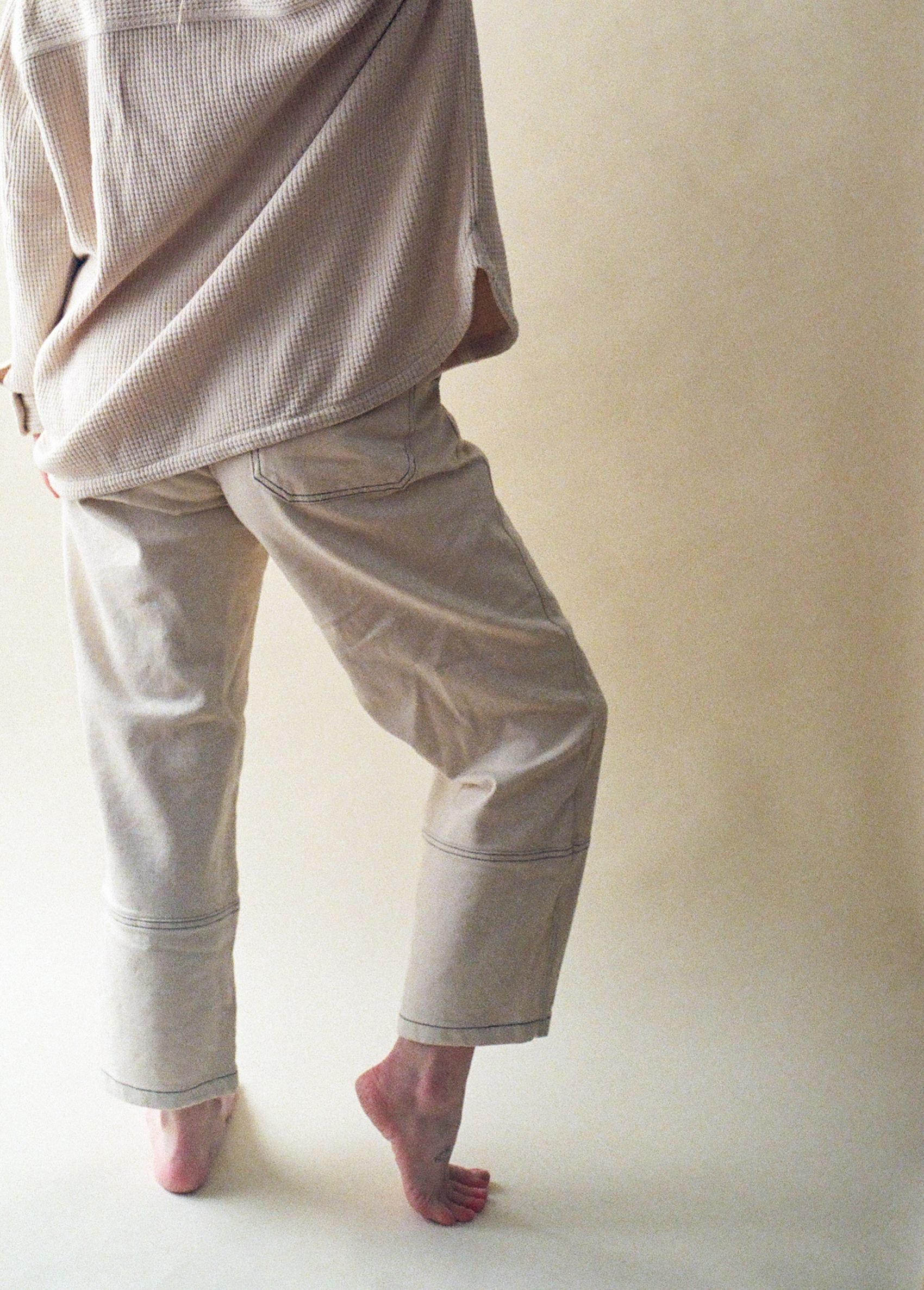 A person standing on one leg, wearing beige pants and a beige top, against a plain beige background.