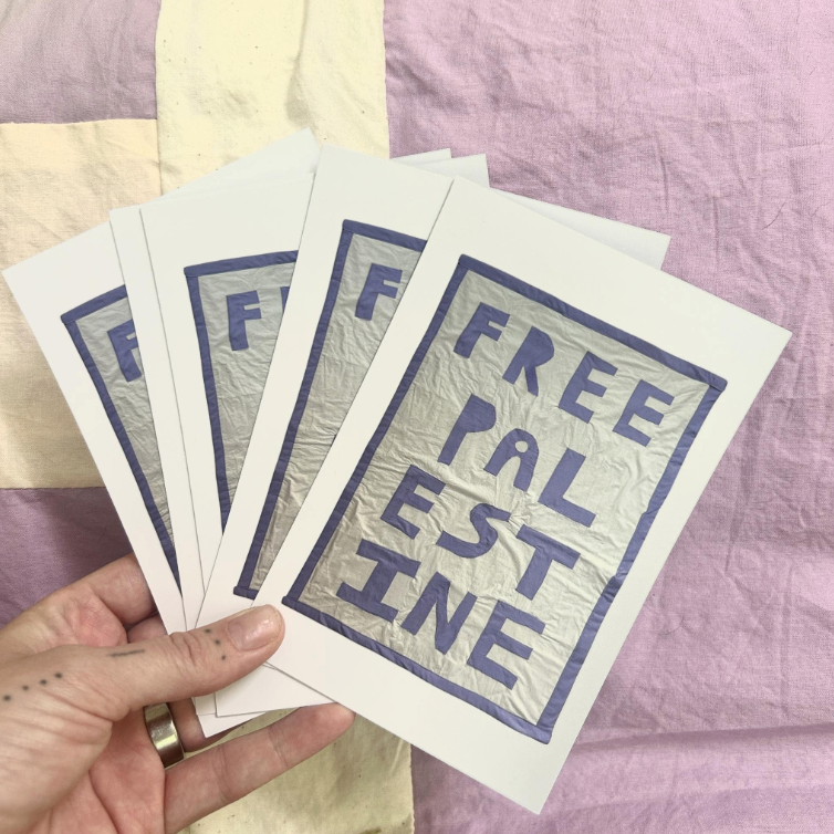 Five printed postcards with the words "FREE PALESTINE" arranged in a square, held in a person's hand against a patchwork fabric background.