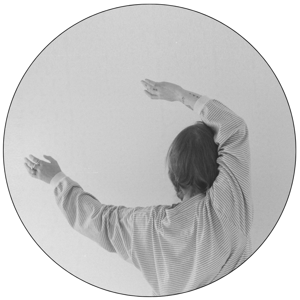 Person with shoulder-length hair wearing a striped long-sleeve shirt, raising arms above head against plain background.