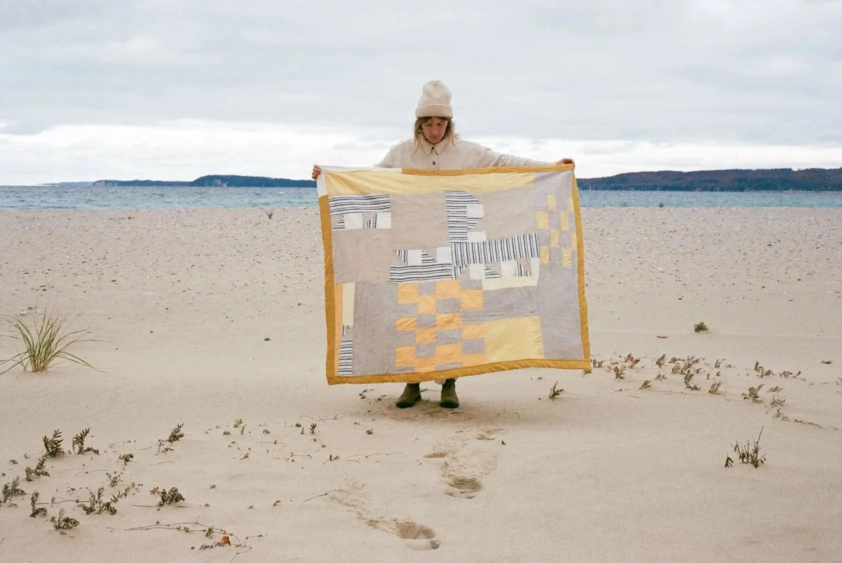 Person standing on a sandy beach holding a quilt with geometric and striped patterns, wearing a beige beanie and coat, with footprints leading to them, and the ocean horizon in the background.