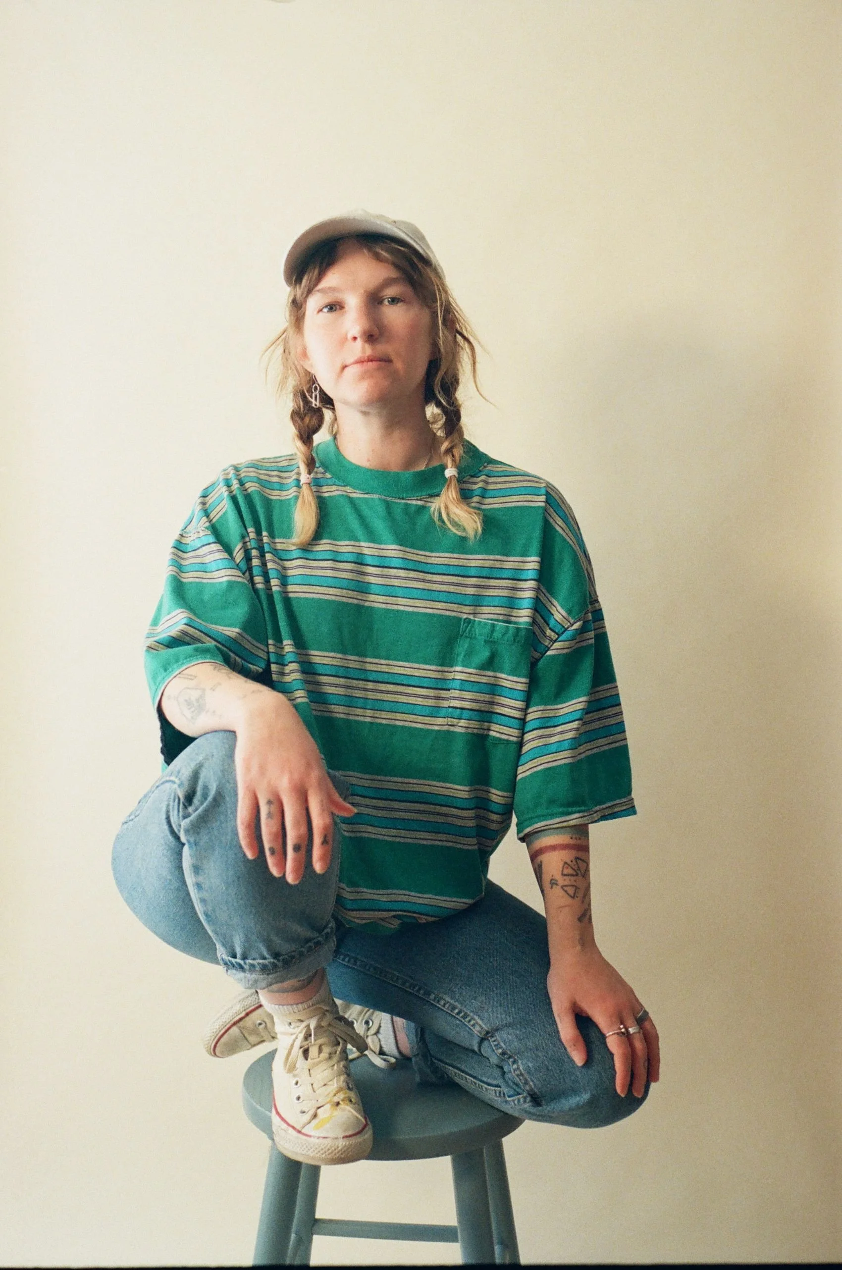 A person with braided hair, wearing a beige cap, green and striped oversized T-shirt, light blue jeans, white sneakers with pink accents, sitting on a stool against a plain off-white wall.