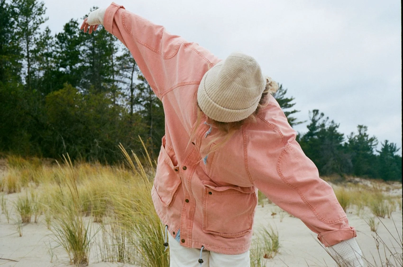 Person with blonde hair wearing a pink jacket, cream knit hat, and white pants standing outdoors in a sandy, grassy area near trees during overcast weather.