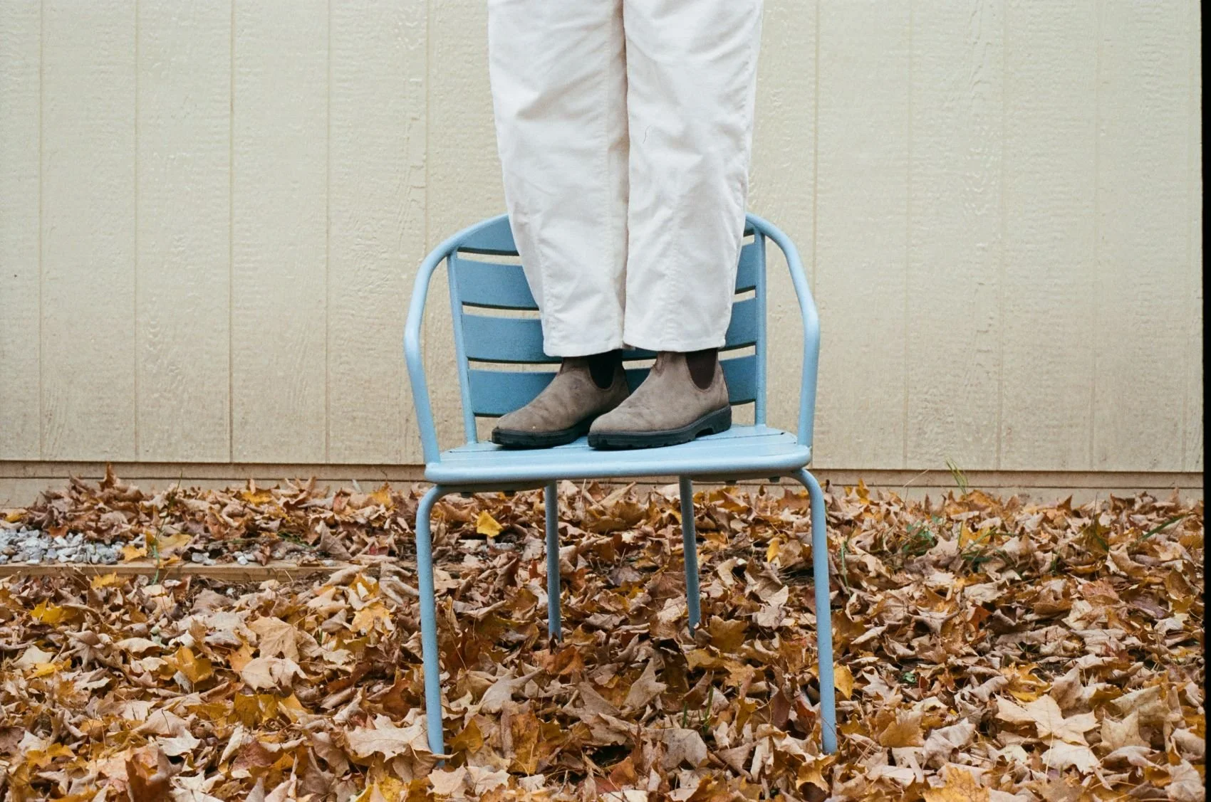 Person standing on a blue metal chair outdoors, with fallen autumn leaves on the ground, wearing light-colored pants and brown boots.